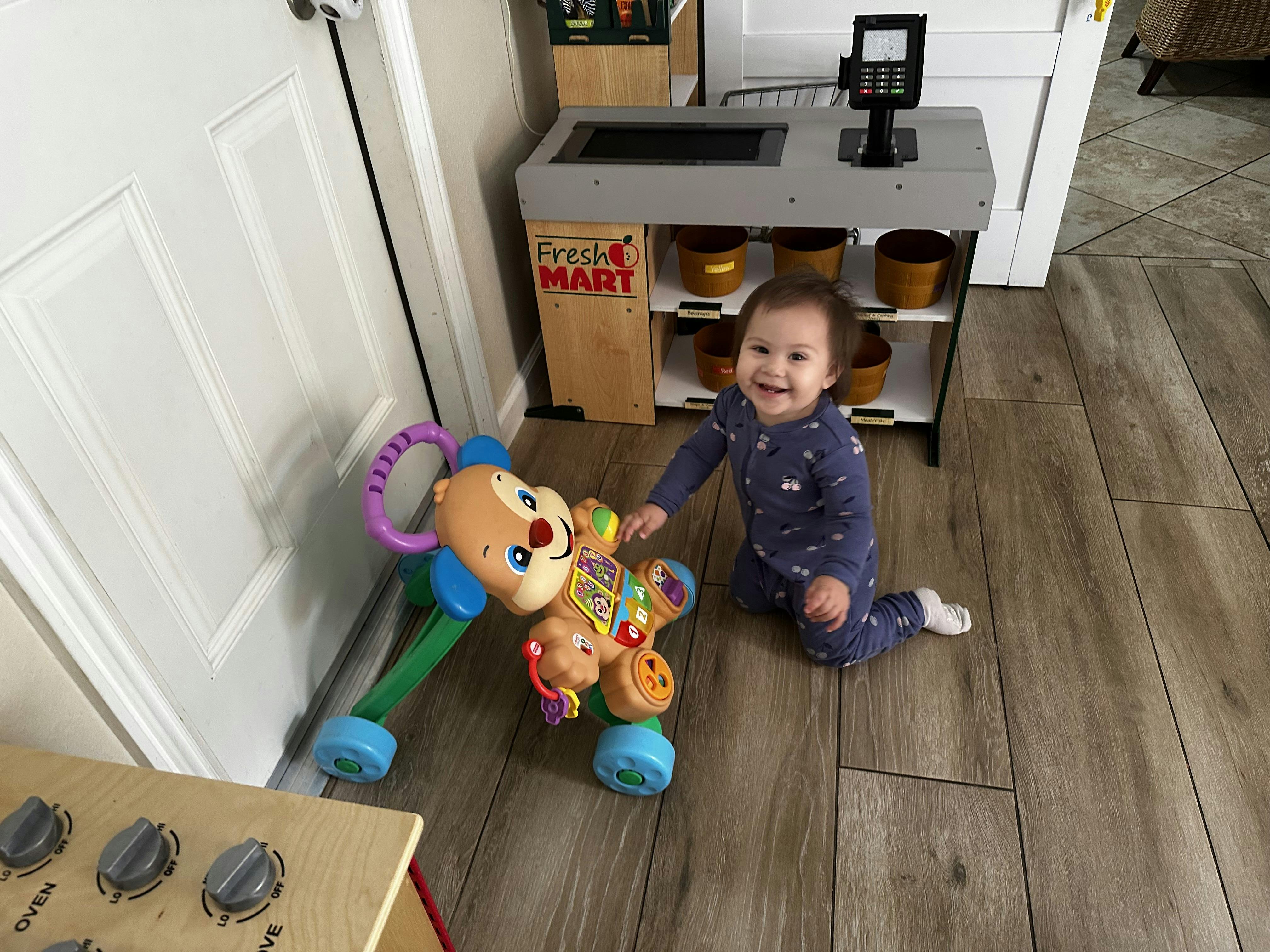 Child on floor next to a Fresh Mart toy stand and a dog-shaped walker toy
