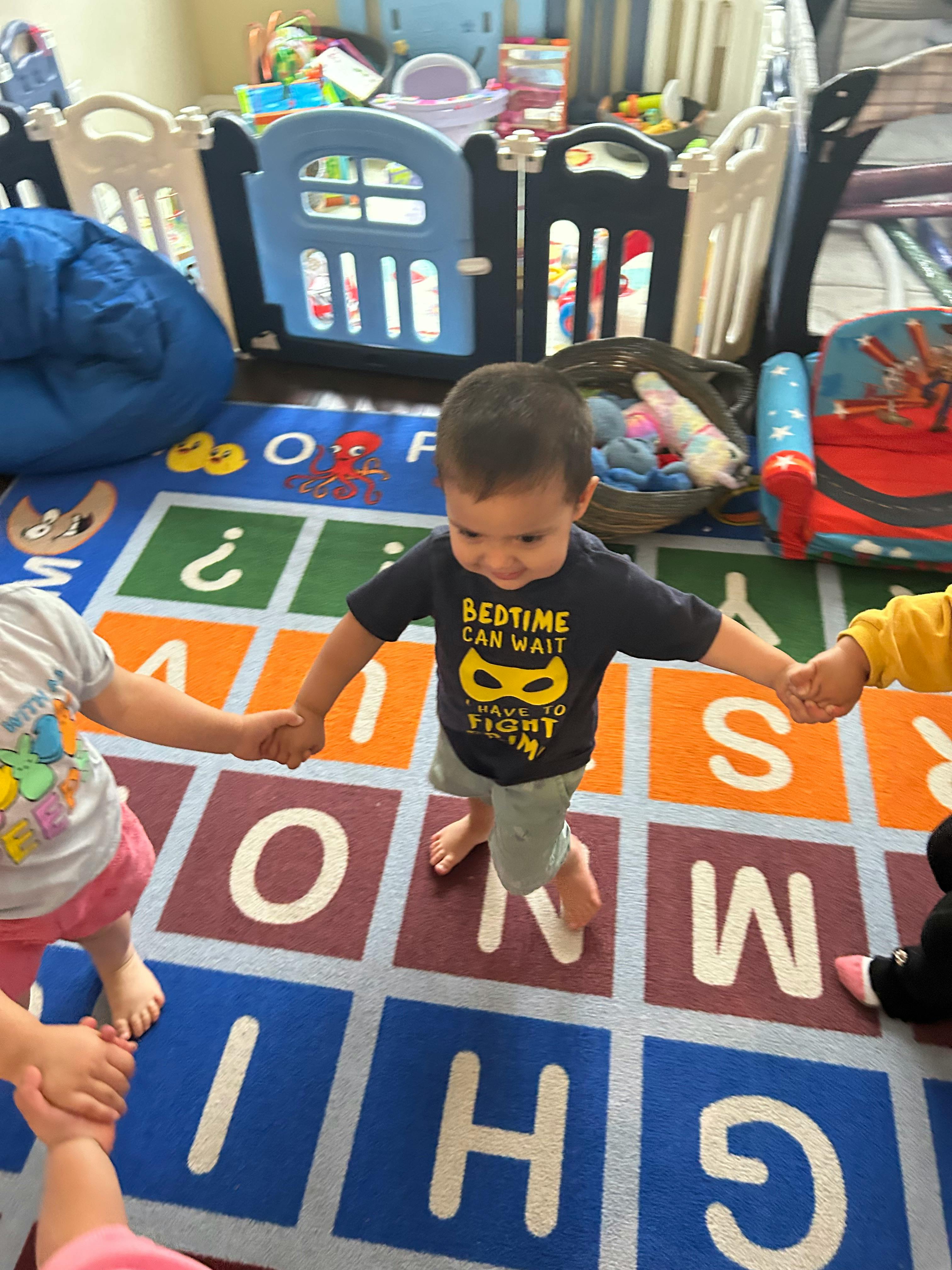 Child in a black shirt holding hands with other children on an alphabet rug