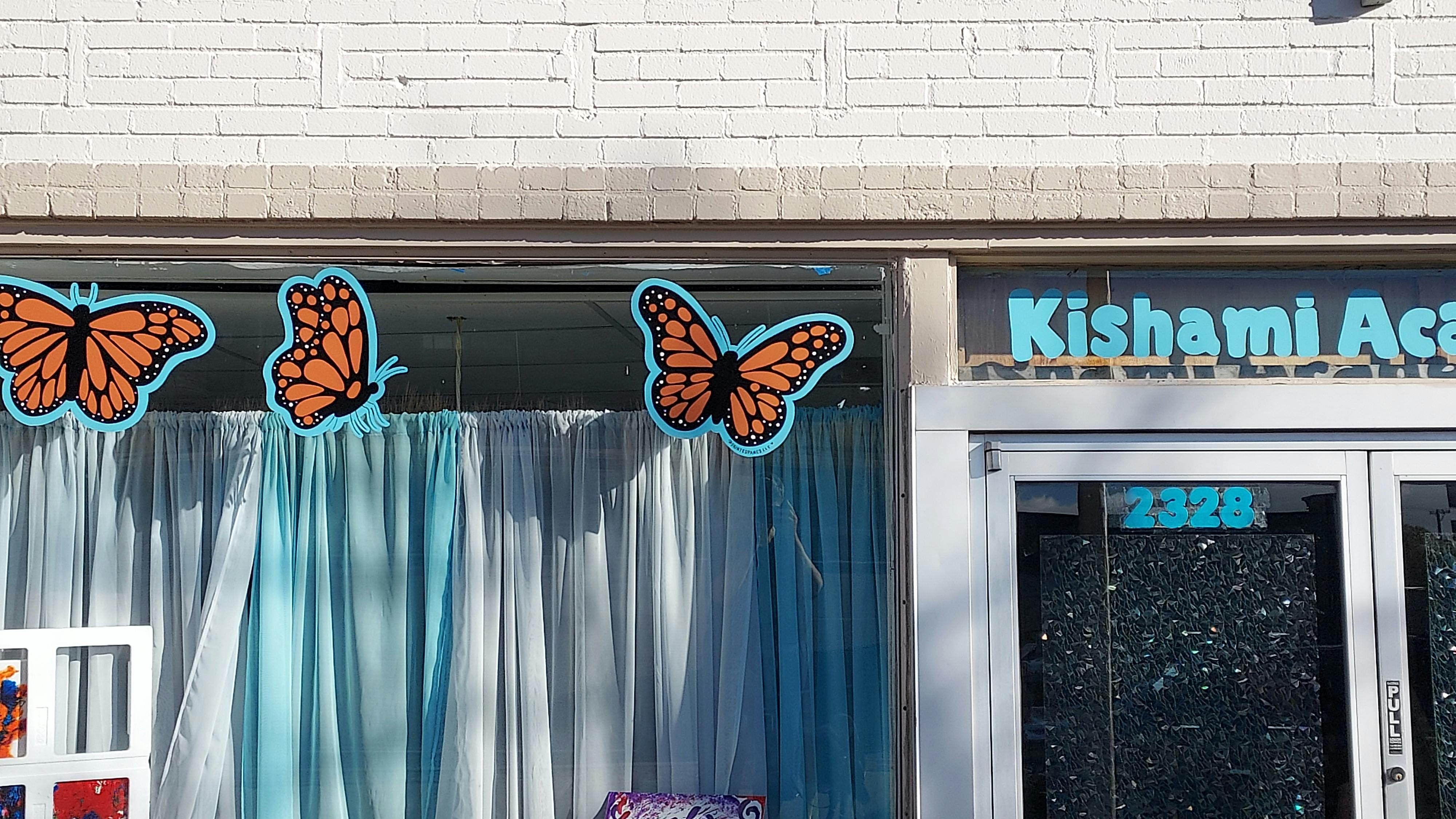 Three butterfly cutouts on a window next to a door with a sign reading "Kishami Academy"