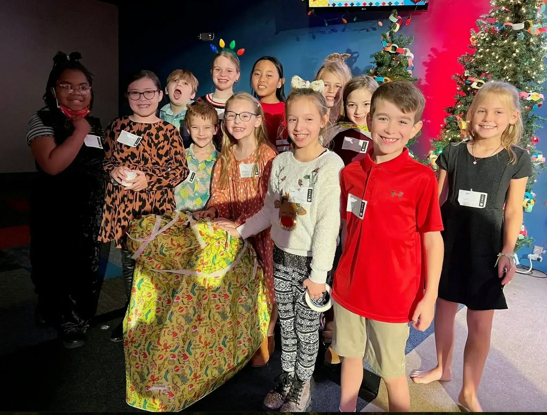 Group of children standing around a large wrapped present, in front of Christmas trees