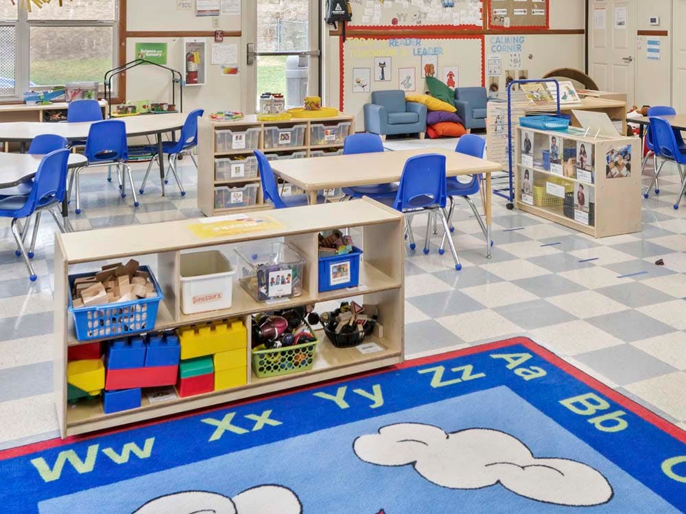 Classroom with tables, blue chairs, shelves with toys, and an alphabet rug