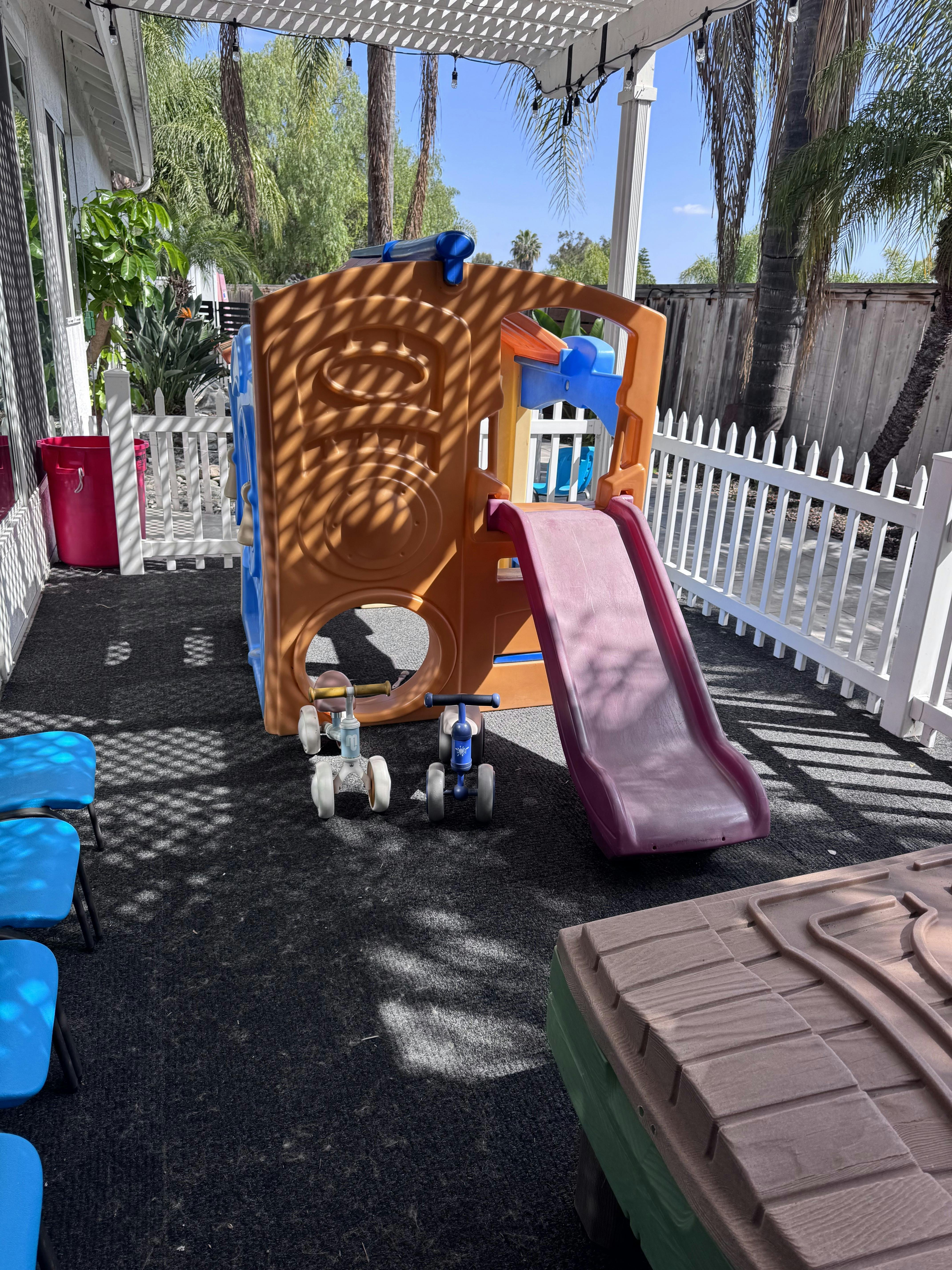 A children's playground structure with a slide, two tricycles, and blue chairs on an outdoor patio