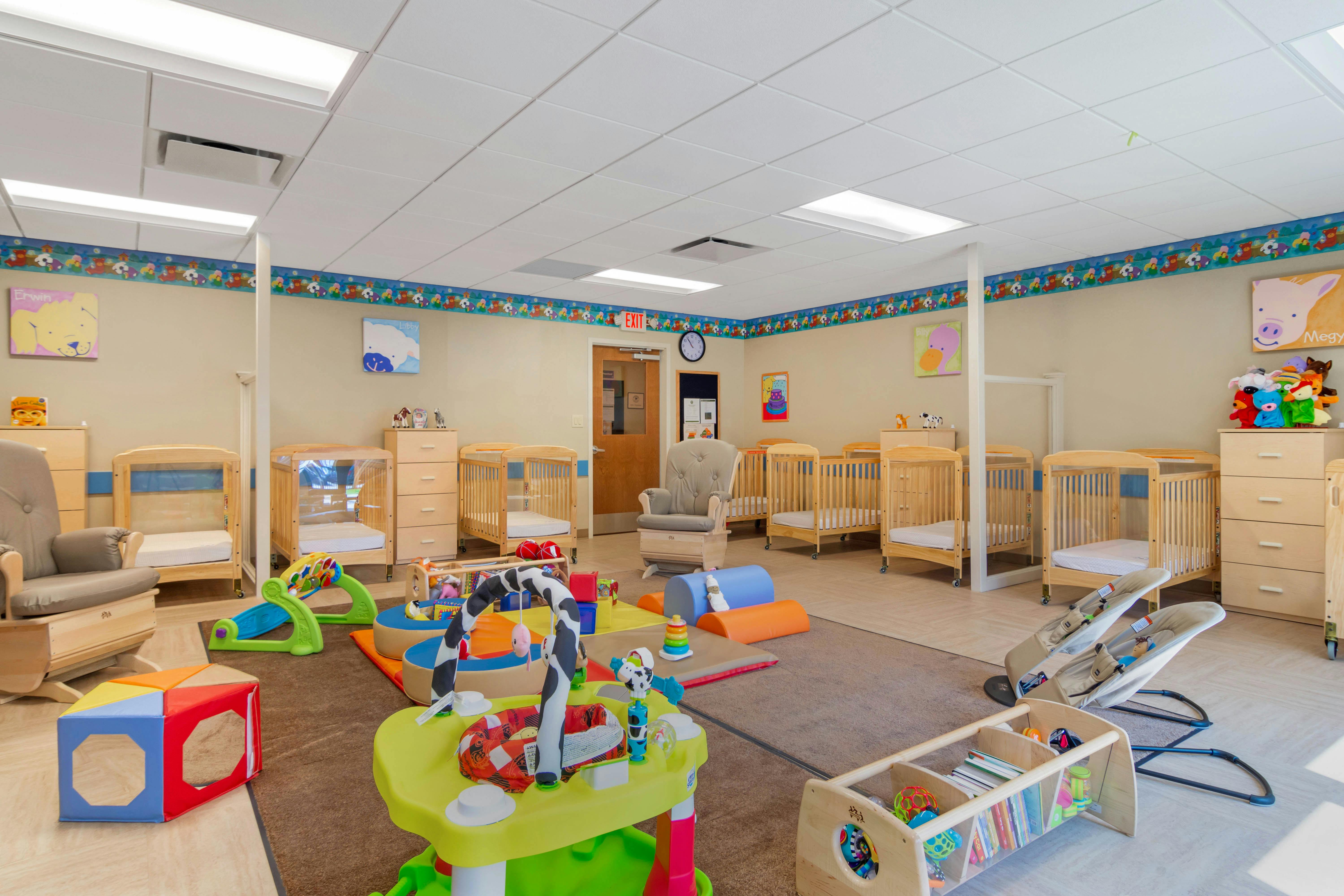 A brightly lit nursery room with multiple cribs, rockers, and various baby toys
