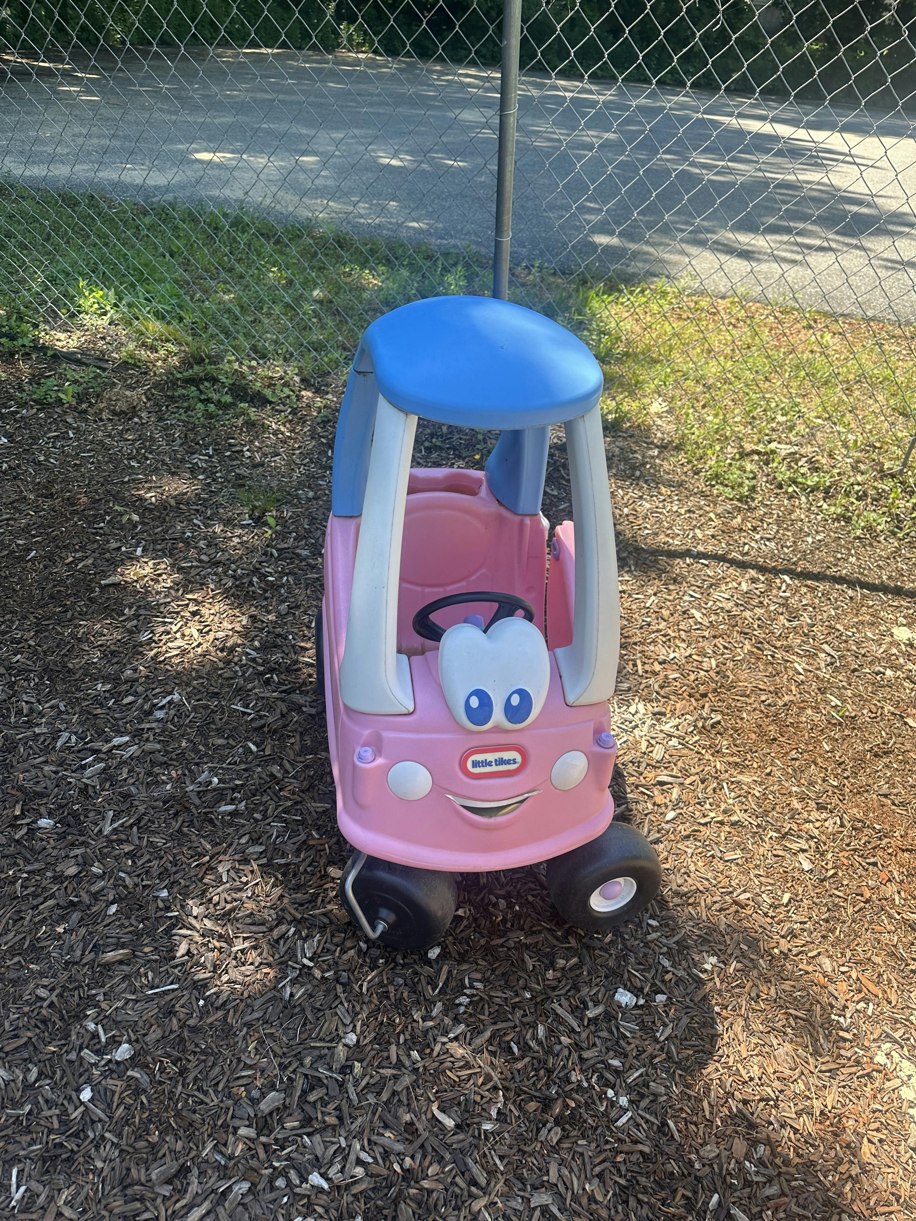 Little Tikes toy car on wood chips by a chain link fence