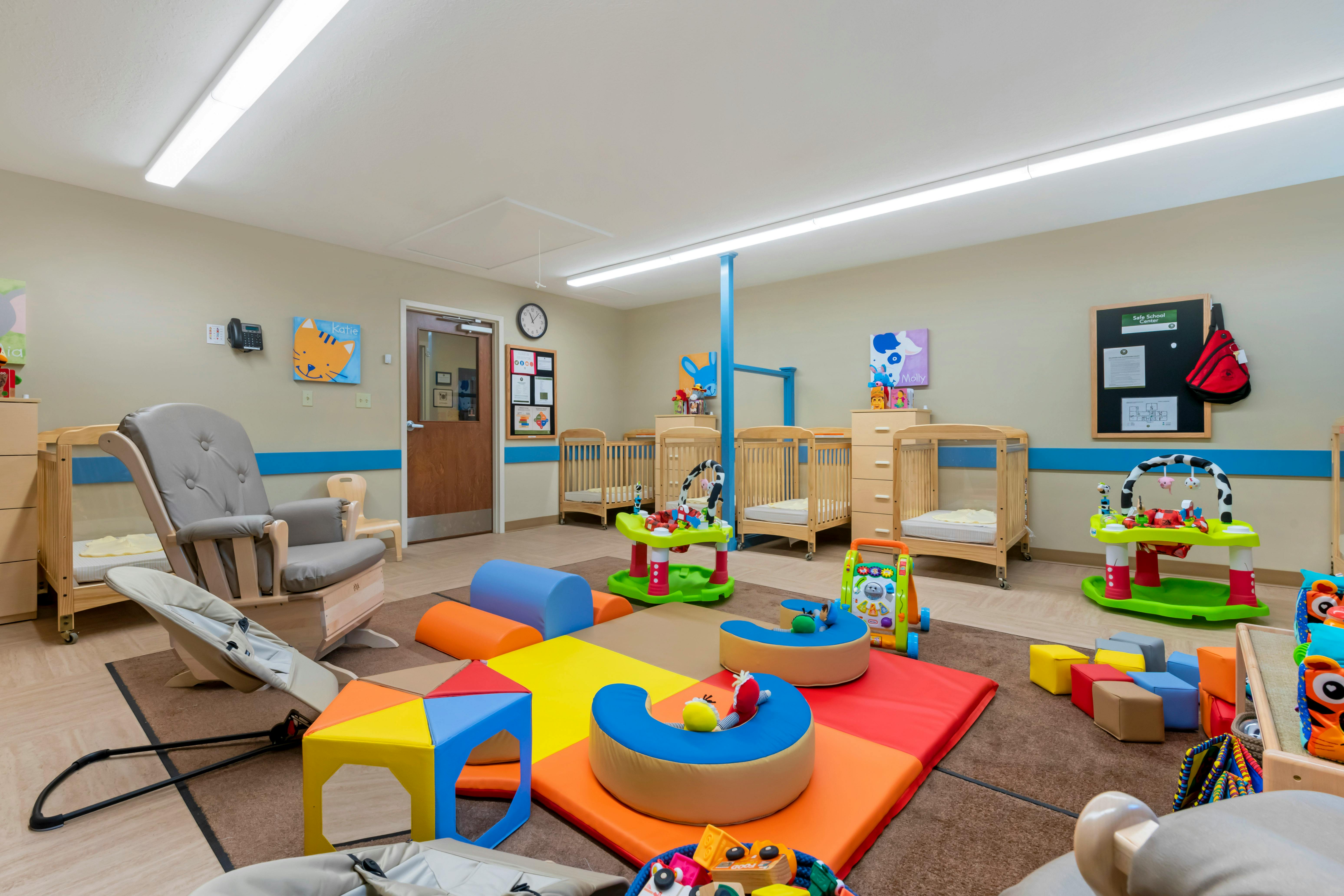 A childcare room with cribs, a glider, and colorful soft play equipment