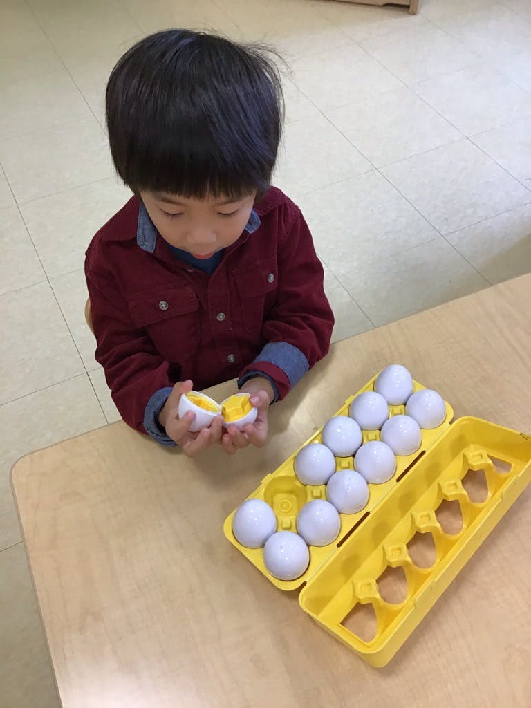 Child holding a toy egg with a yellow yolk, next to an egg carton with toy eggs