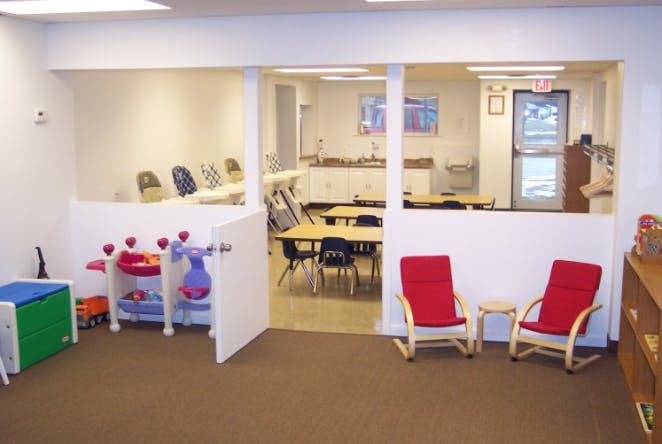 Playroom with toys and two red chairs, classroom with tables and chairs beyond