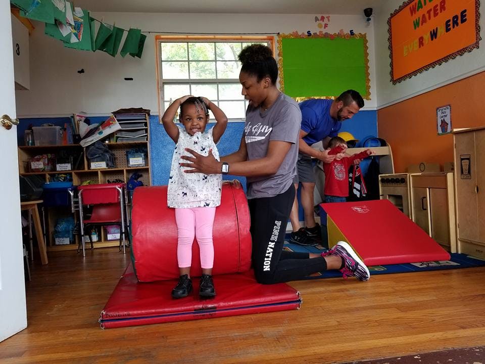 Adult helping a child stand on a red cylinder in a classroom