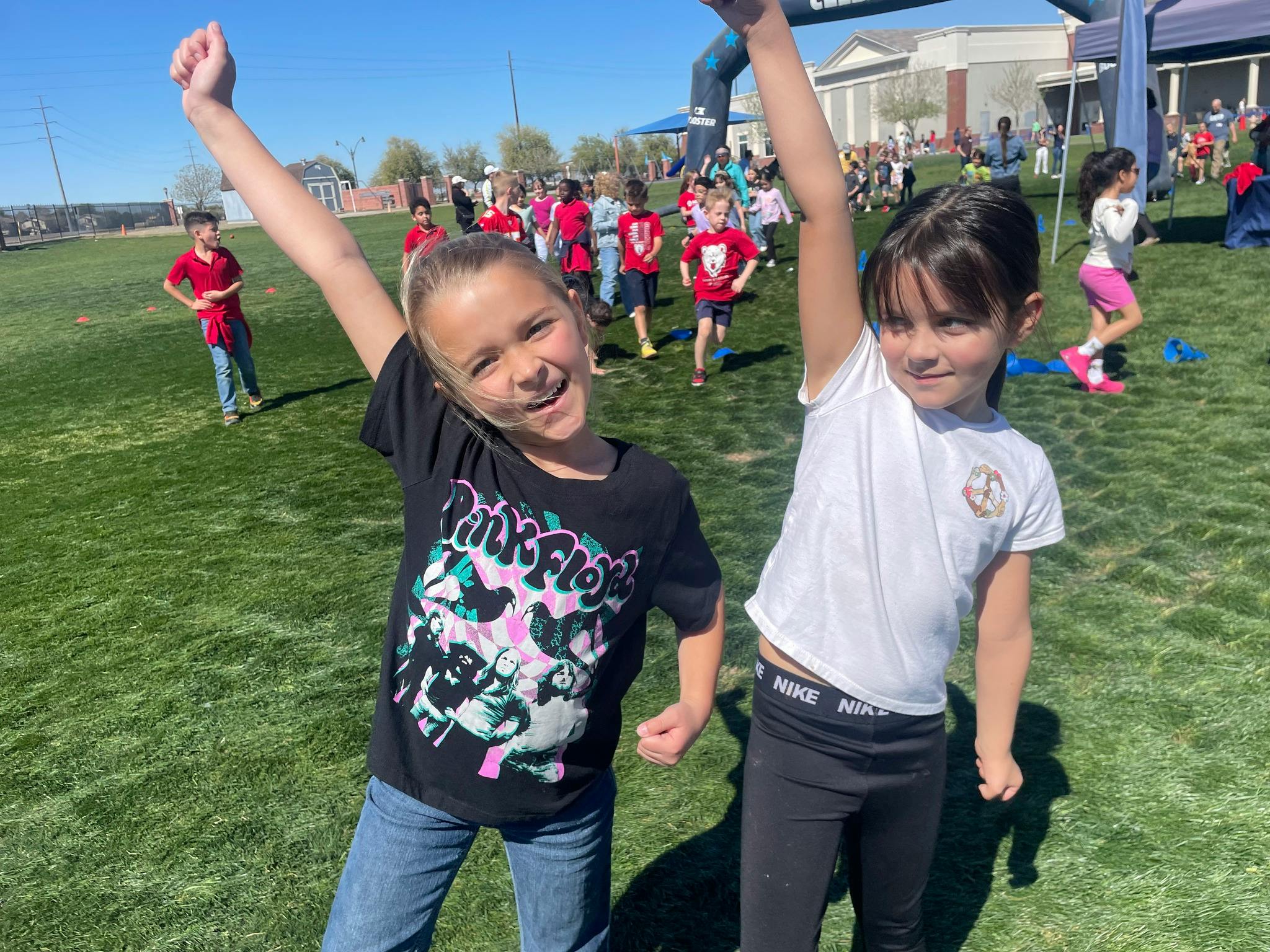 Two children with arms raised in a field with other people in the background