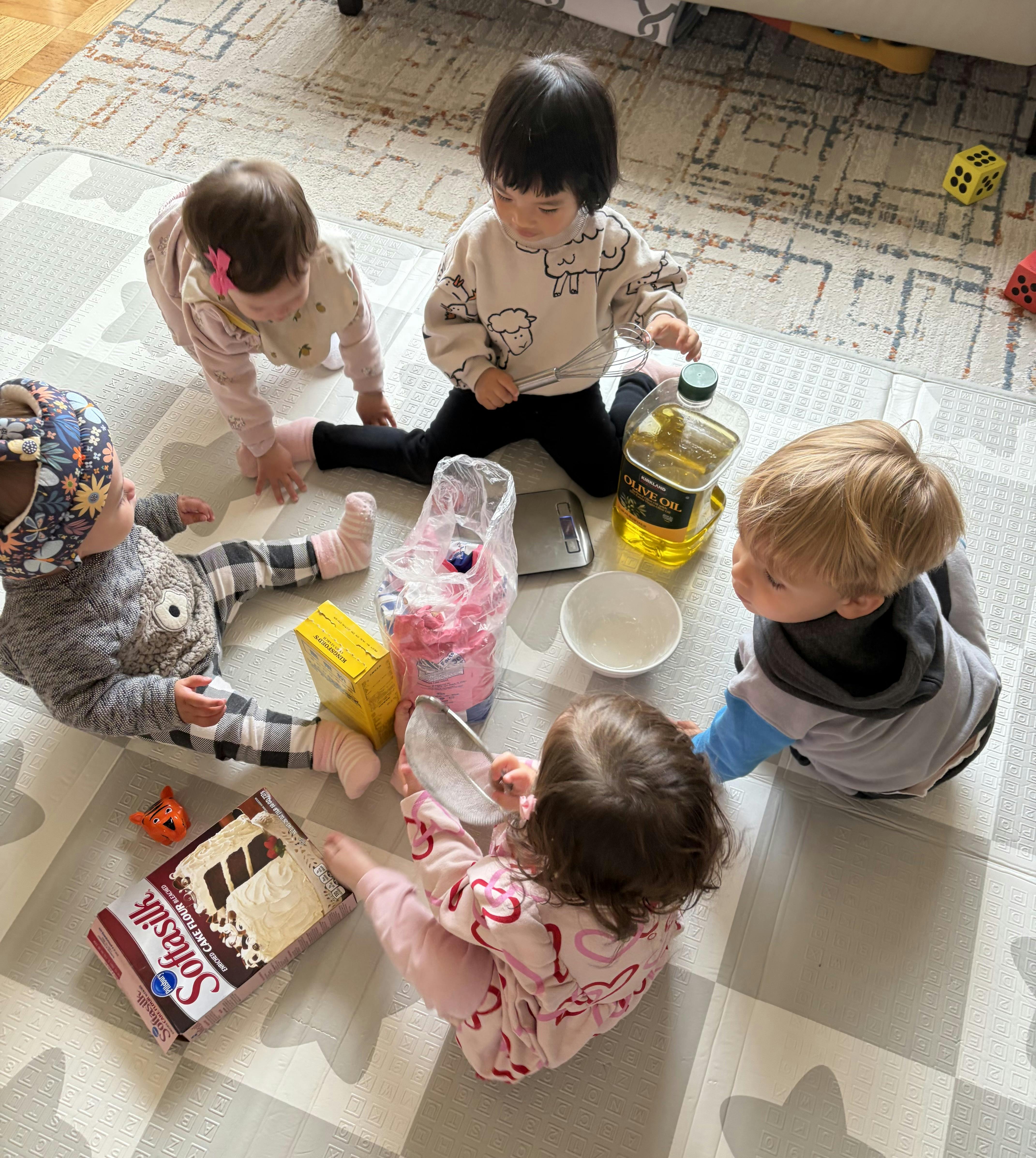 Five children sitting on a play mat with baking supplies