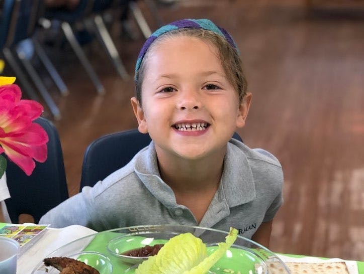Child wearing a kippah, smiling at a table with food