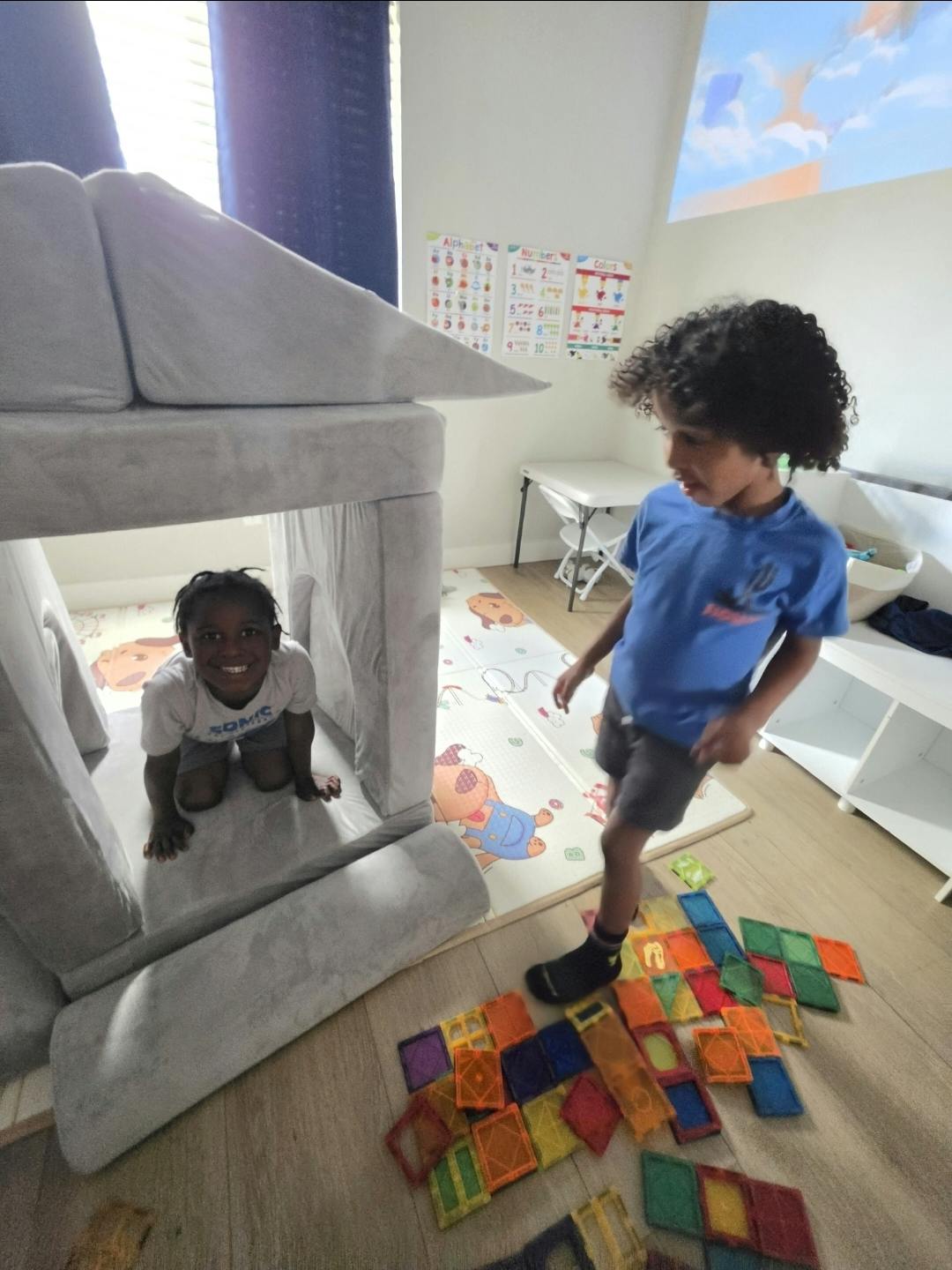 Child peeking from a play fort as another child stands next to magnetic tiles