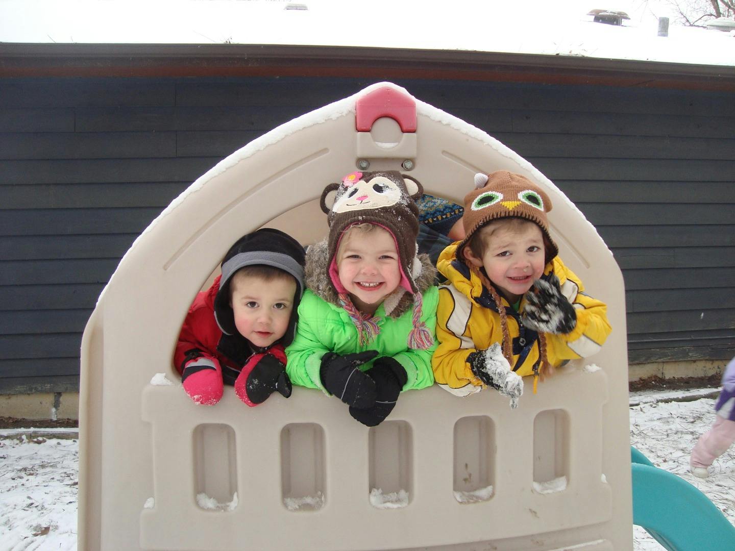 Three children in winter clothing looking out from a playhouse