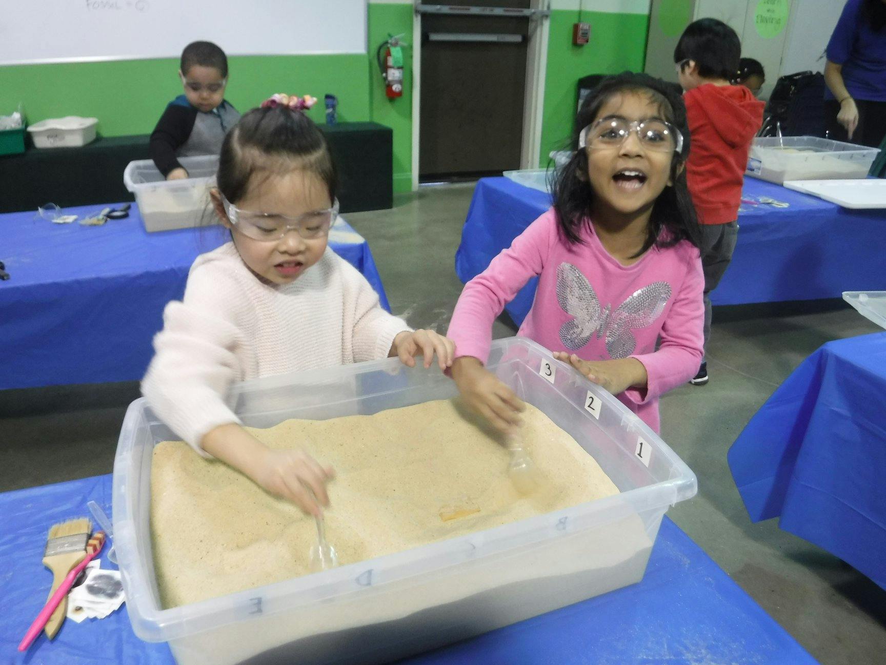 Two children wearing safety glasses playing with sand in a container