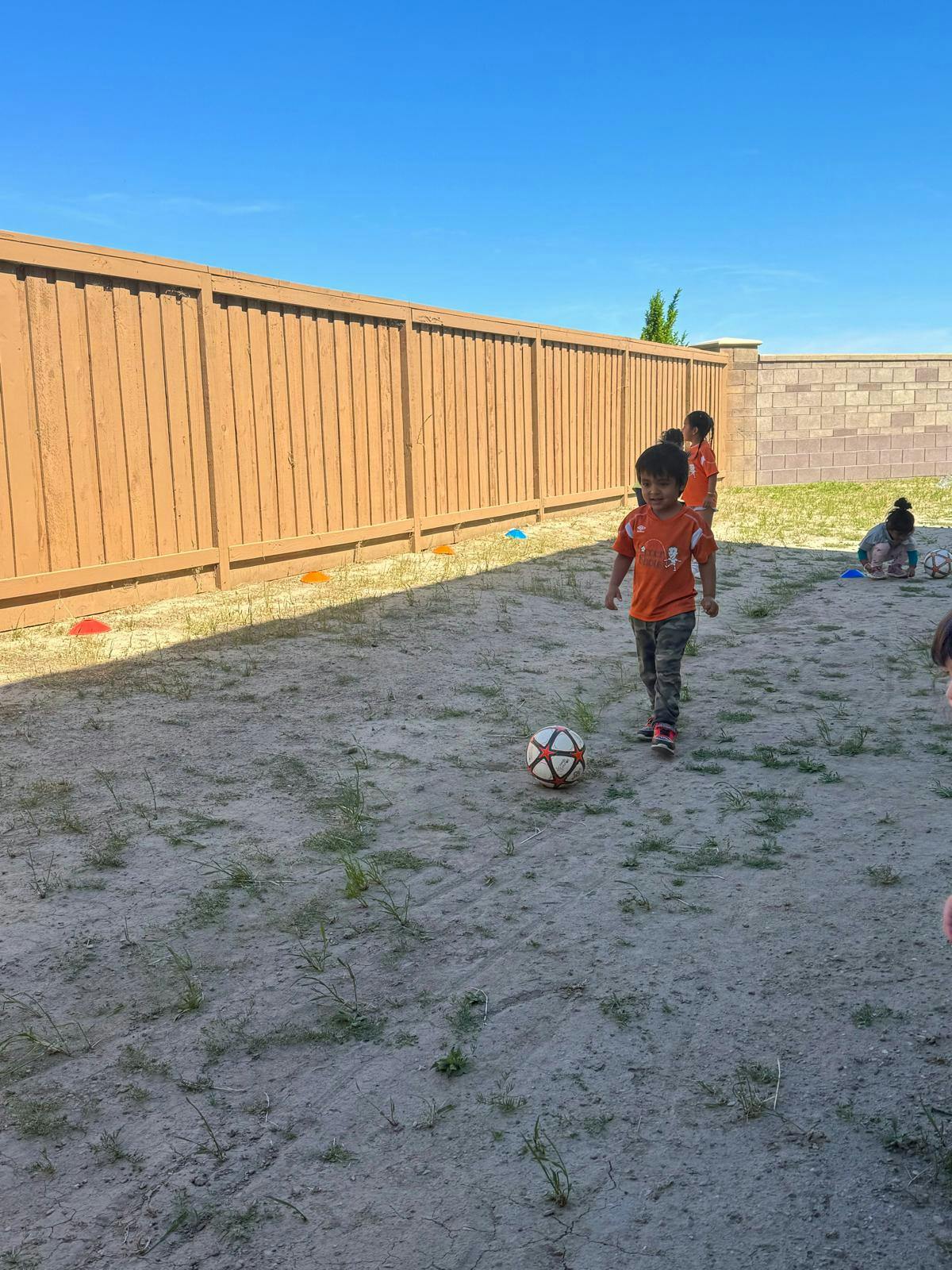 Child in orange shirt standing near a soccer ball on dirt