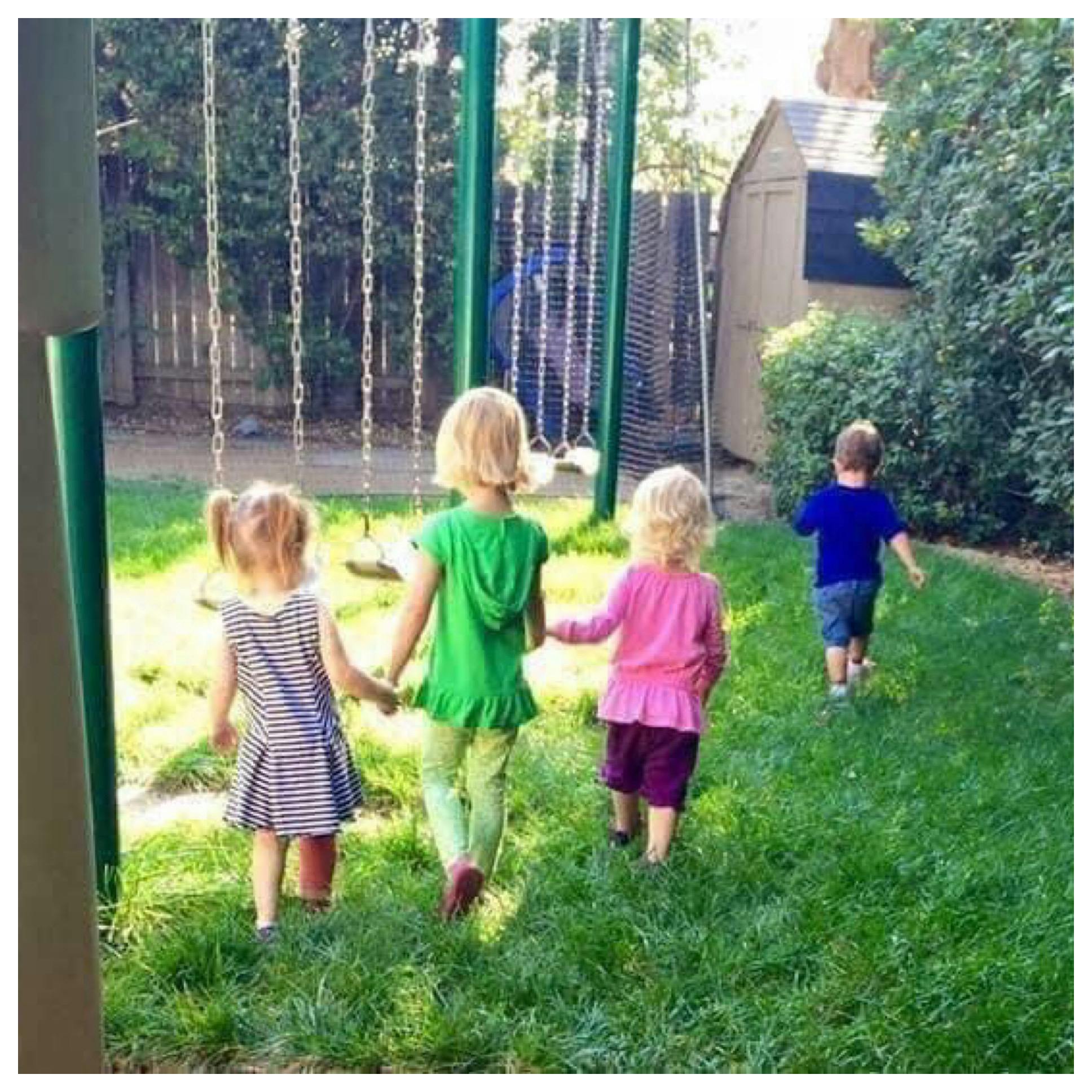 Three children holding hands walking toward a swing set, another child walking away