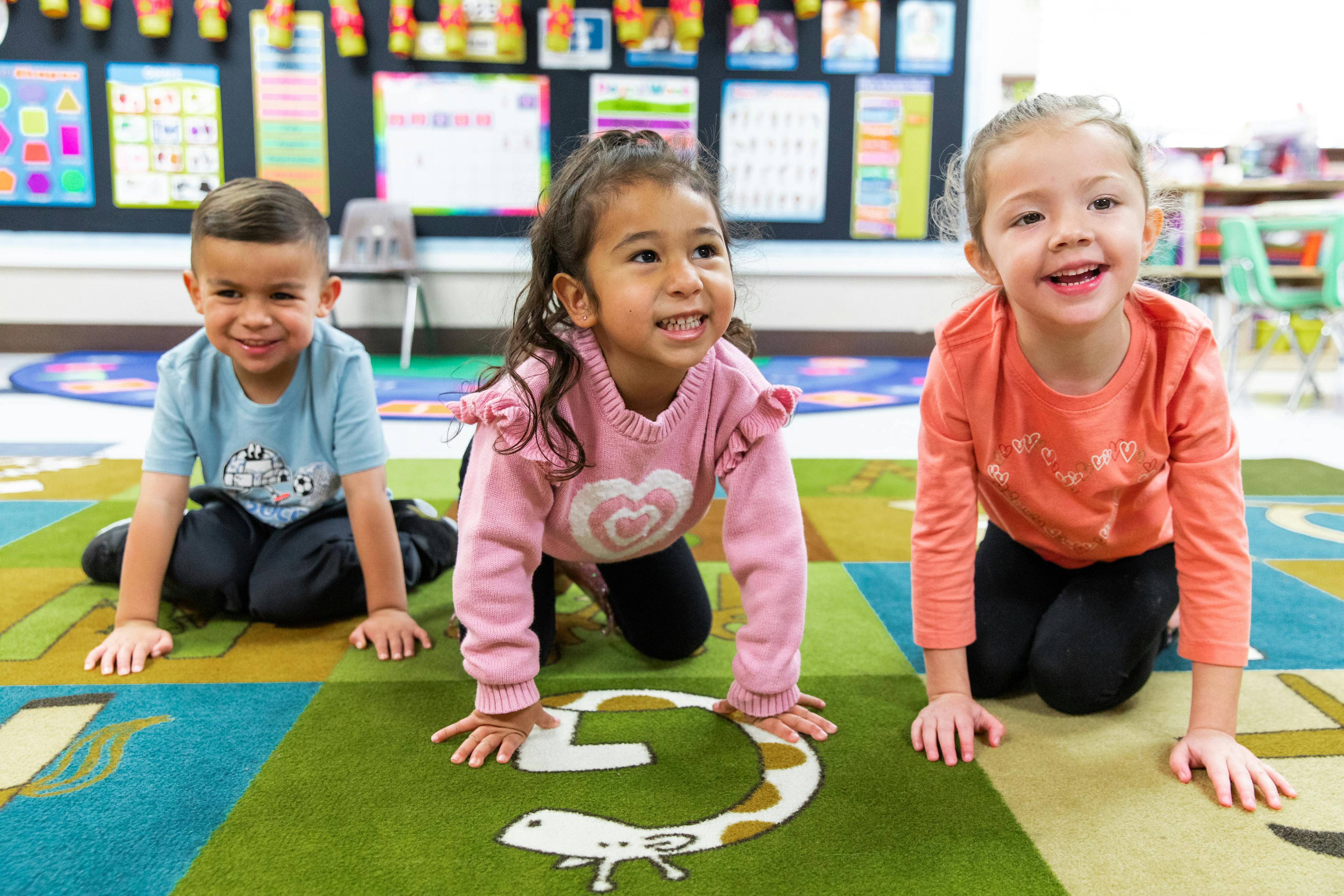 Three children on hands and knees on a colorful rug in a classroom