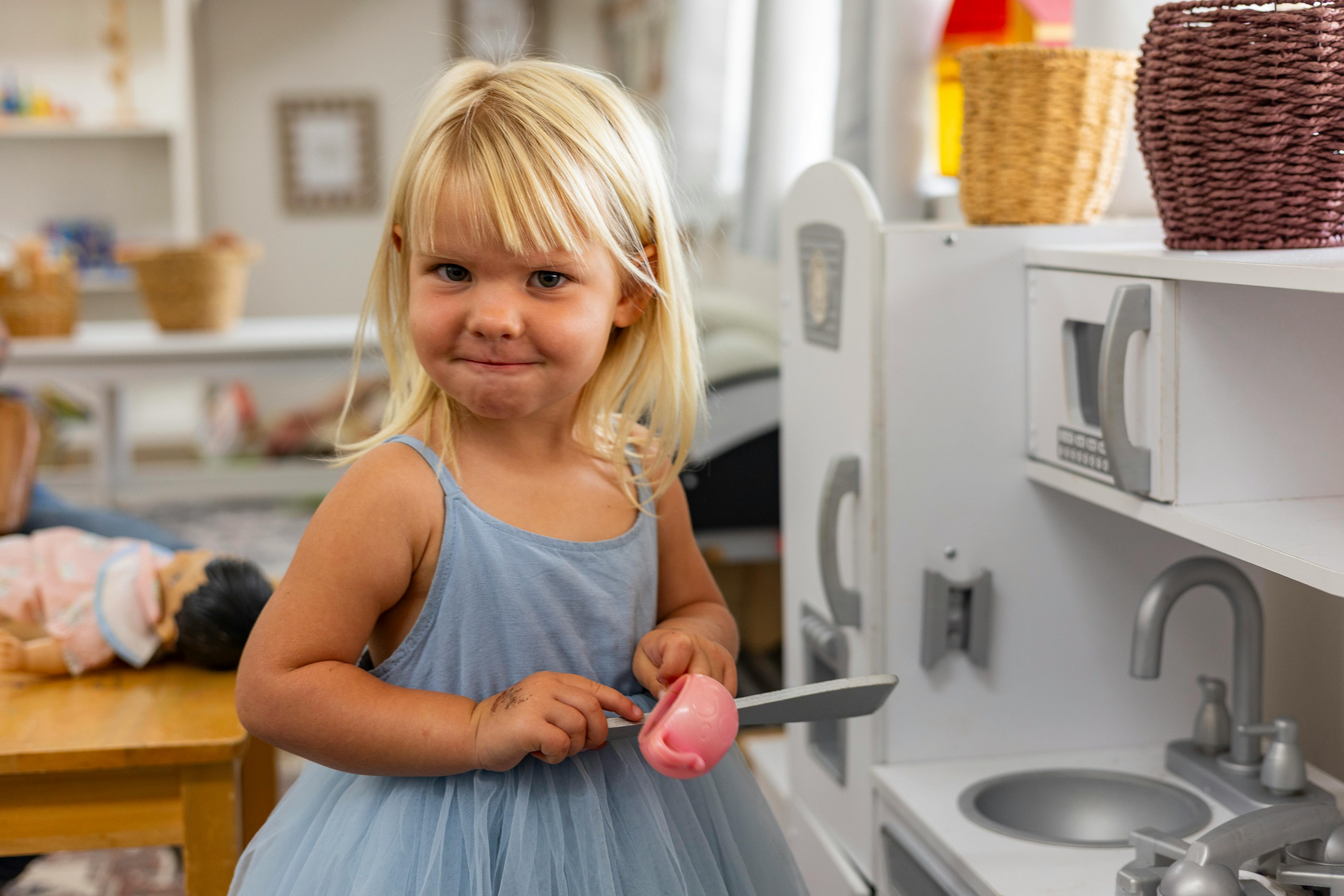 Child in blue dress holding toy food and knife next to toy kitchen
