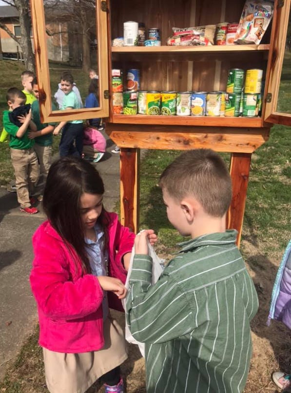 Two children interacting in front of a small outdoor pantry filled with canned goods