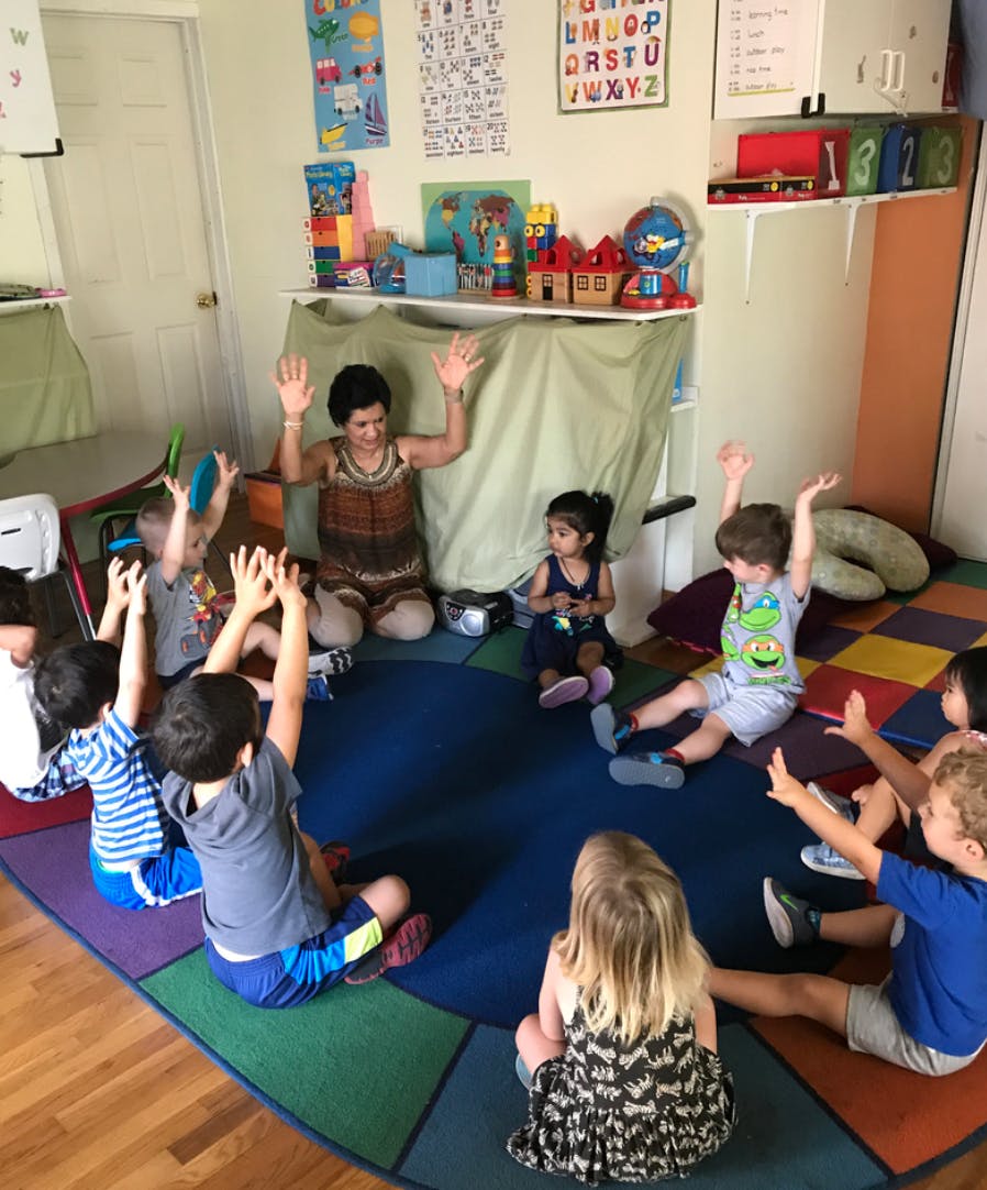 Adult and children sitting in a circle with hands raised in a classroom