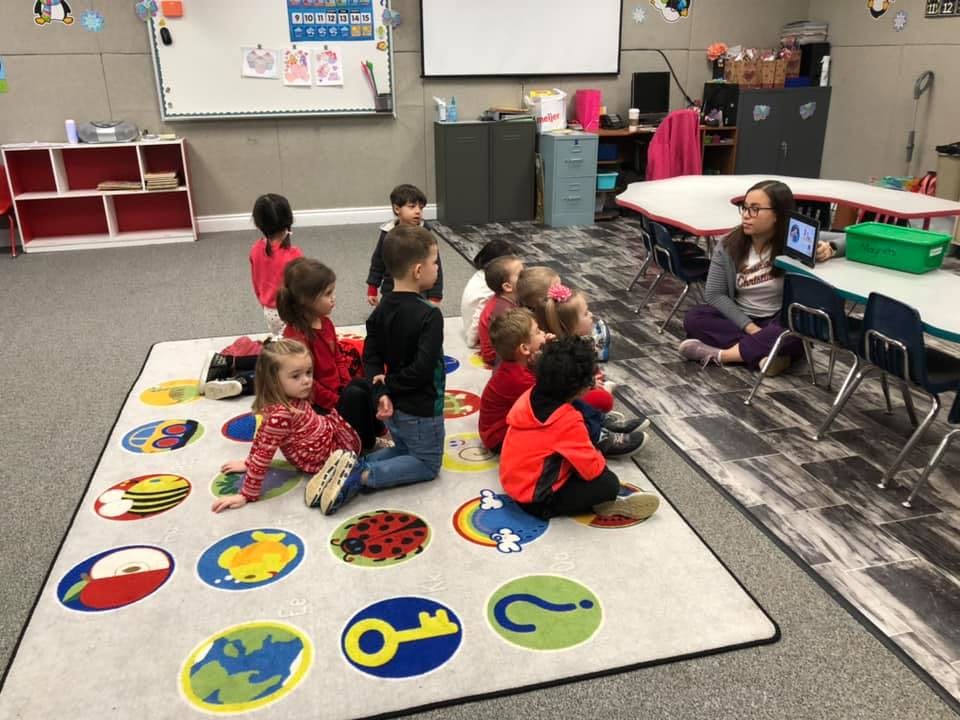 Children sitting on a rug with a person sitting at a table