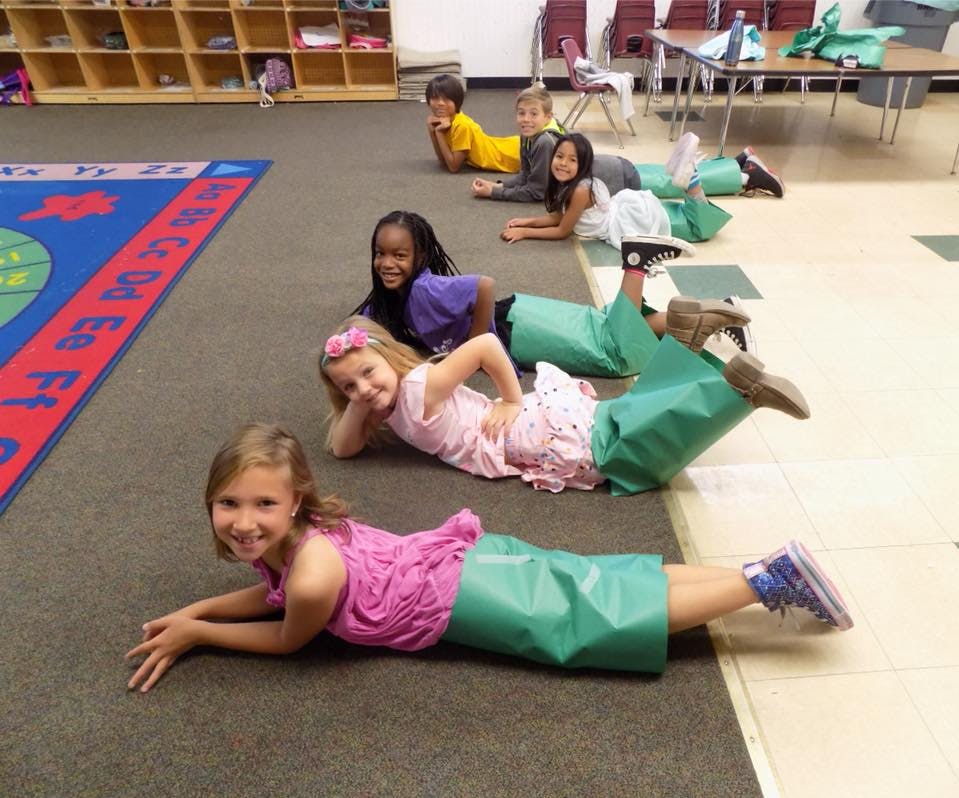 Children lying on the floor in a classroom, some with green bags on their legs
