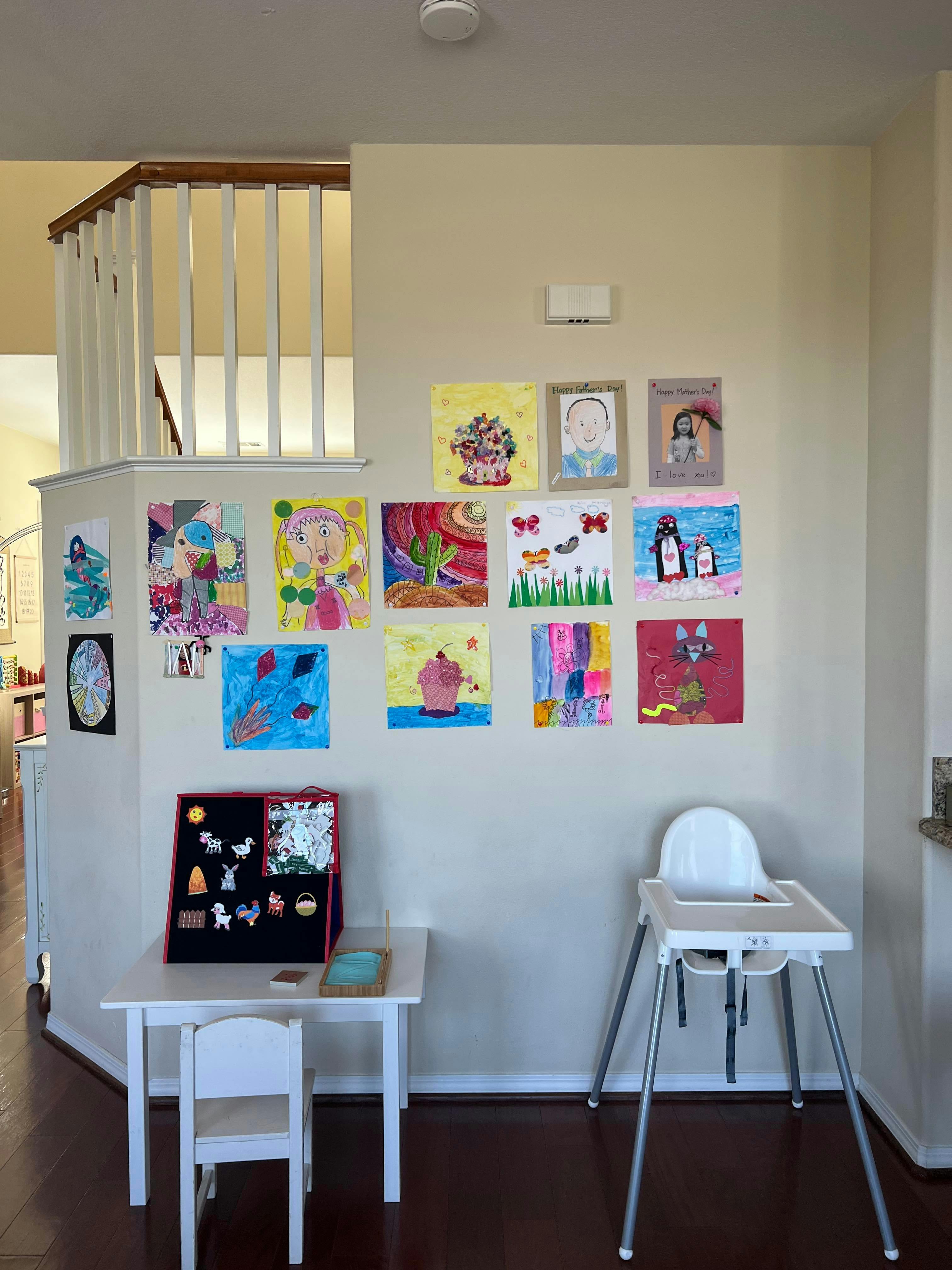 Children's artwork displayed on a wall above a small table and high chair