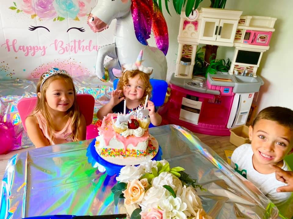 Three children celebrating a birthday with a cake, unicorn balloon, and Happy Birthday banner