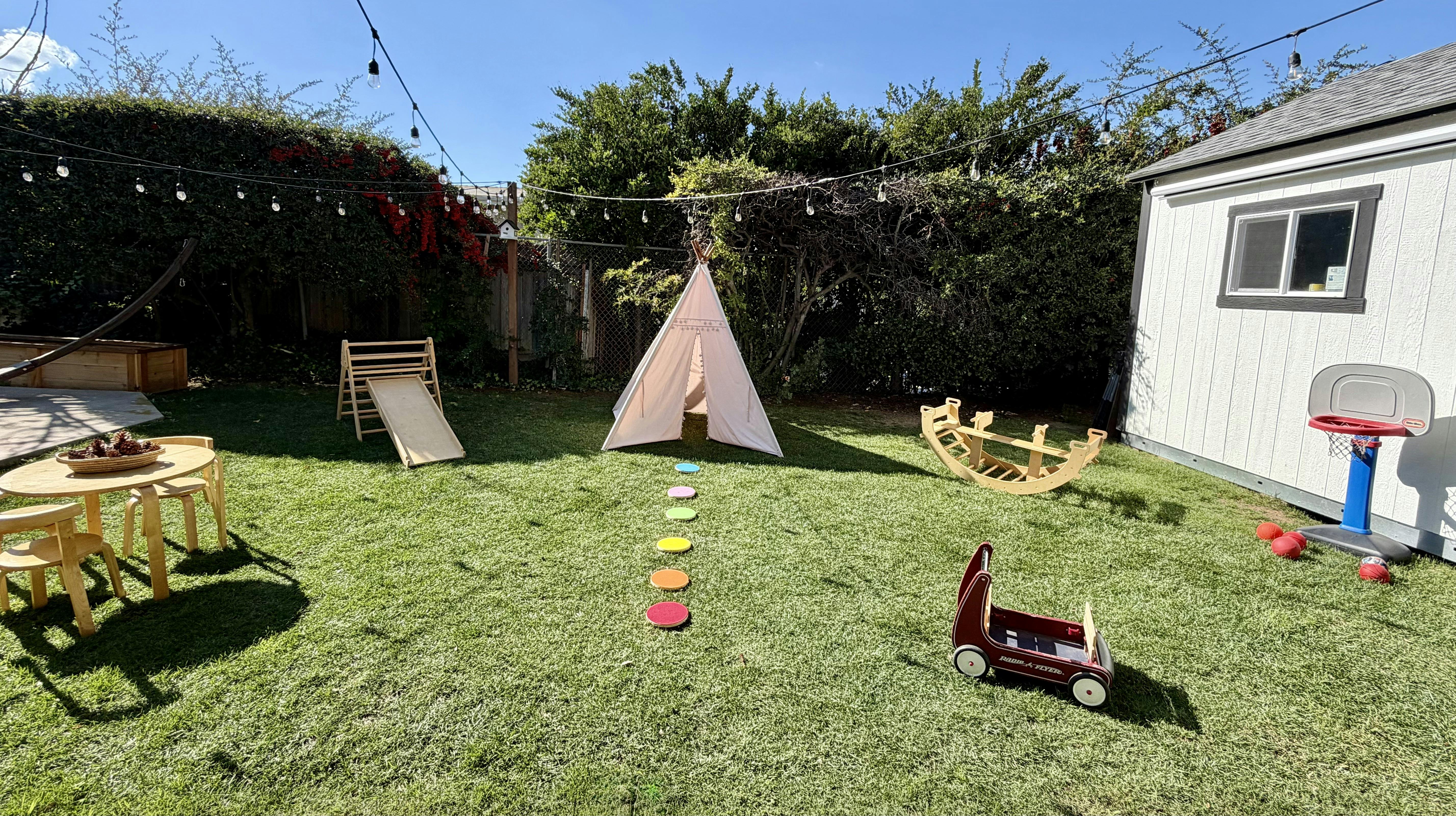 Backyard with a small slide, teepee, toy wagon, wooden rocking toy, and basketball hoop