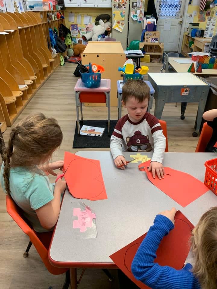 Children cutting red paper at a table in a classroom