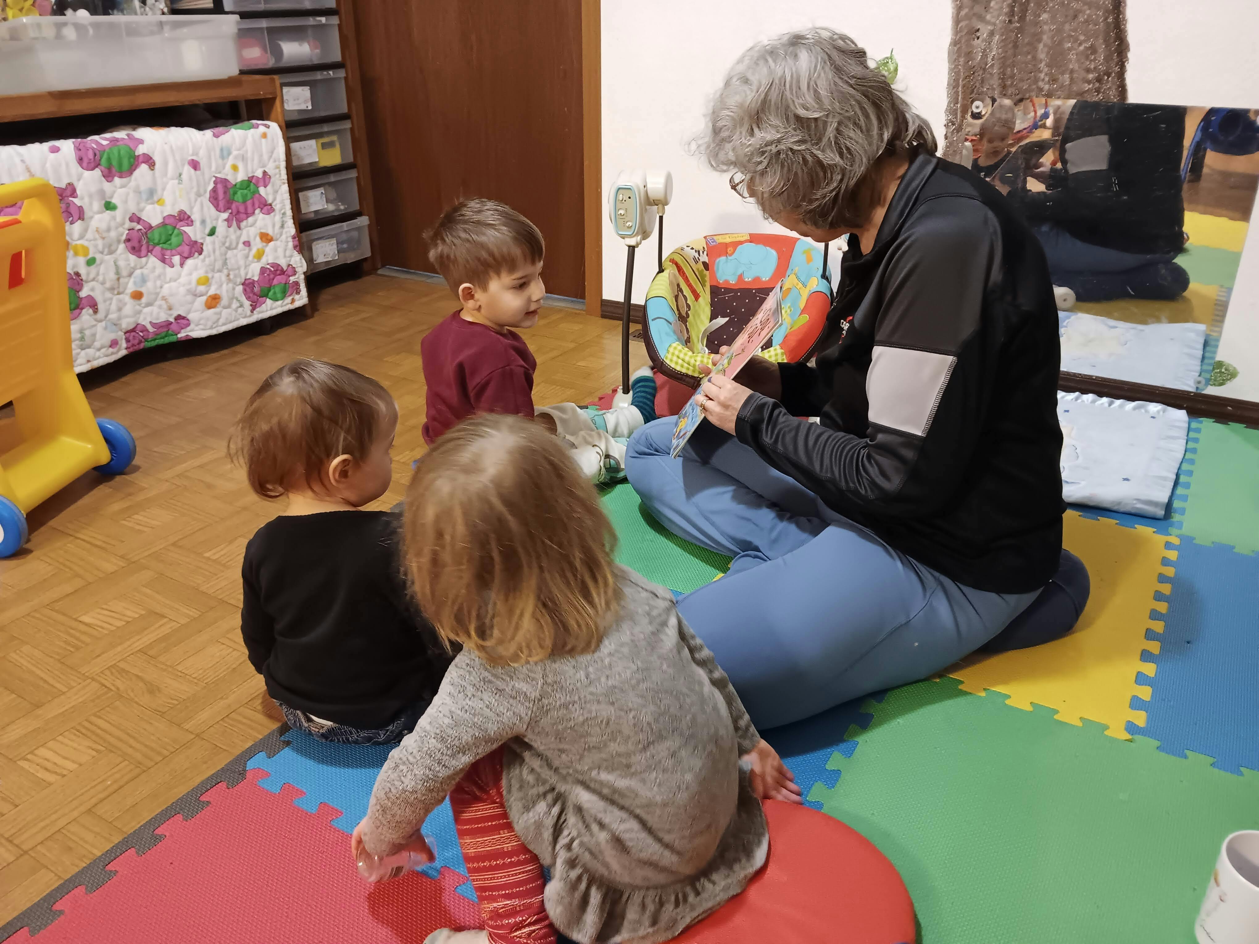 Adult reading a book to three children sitting on a play mat