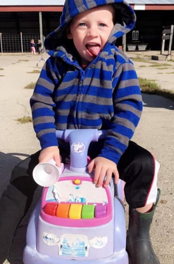 Child in a striped hoodie sitting on a toy car with tongue out