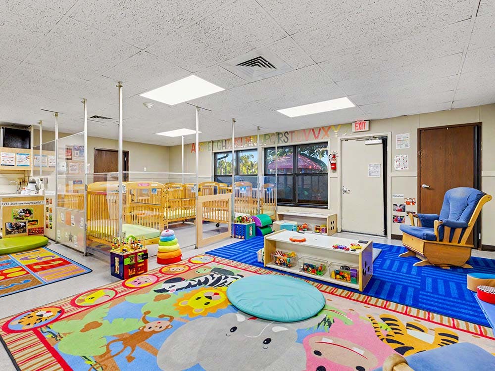 A colorful childcare room with cribs, playmats, and a rocking chair