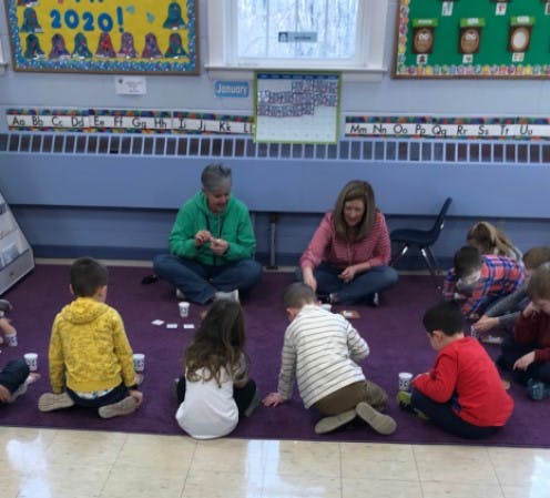 Two adults and multiple children sitting on a purple rug in a classroom