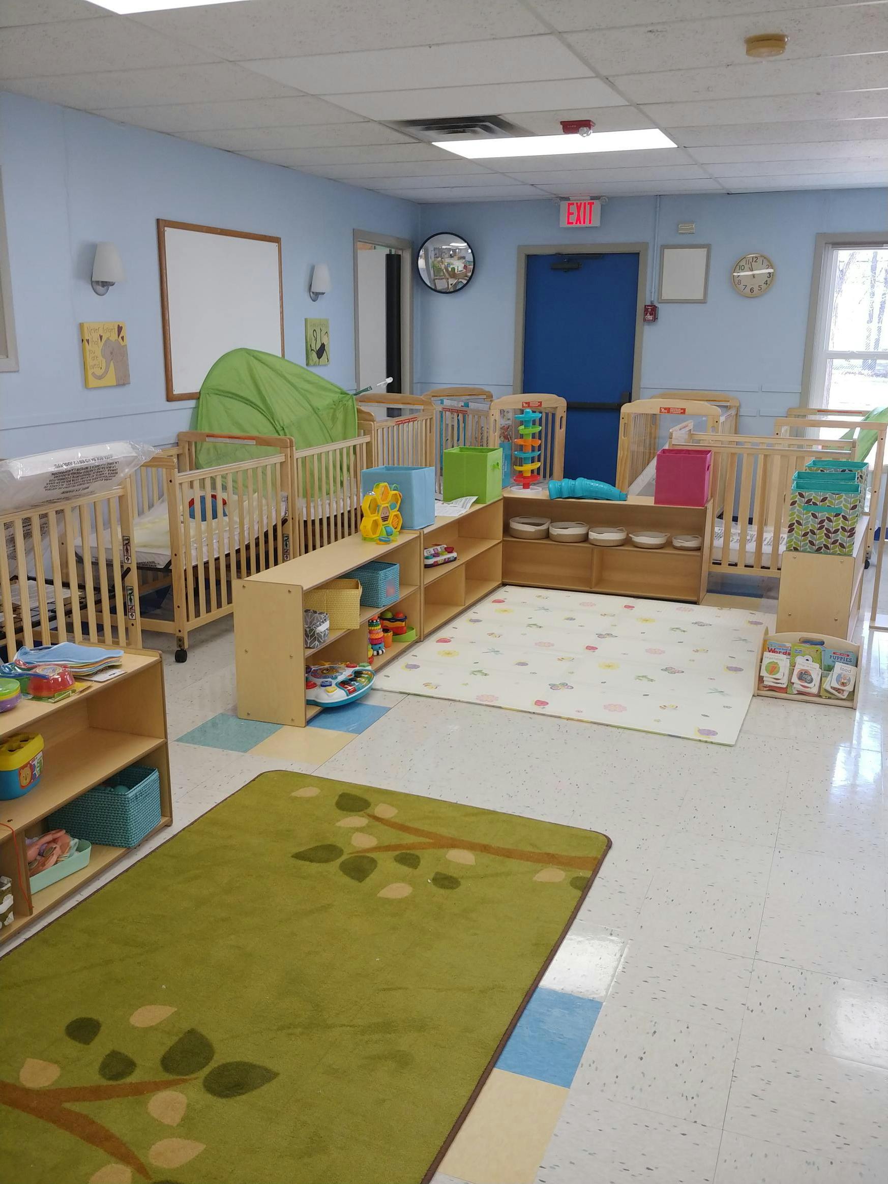 Nursery room with multiple cribs, shelves with toys, and two rugs on the floor