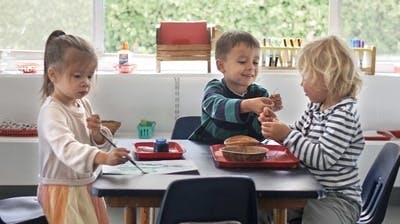Three children at a table, one painting and two interacting with objects