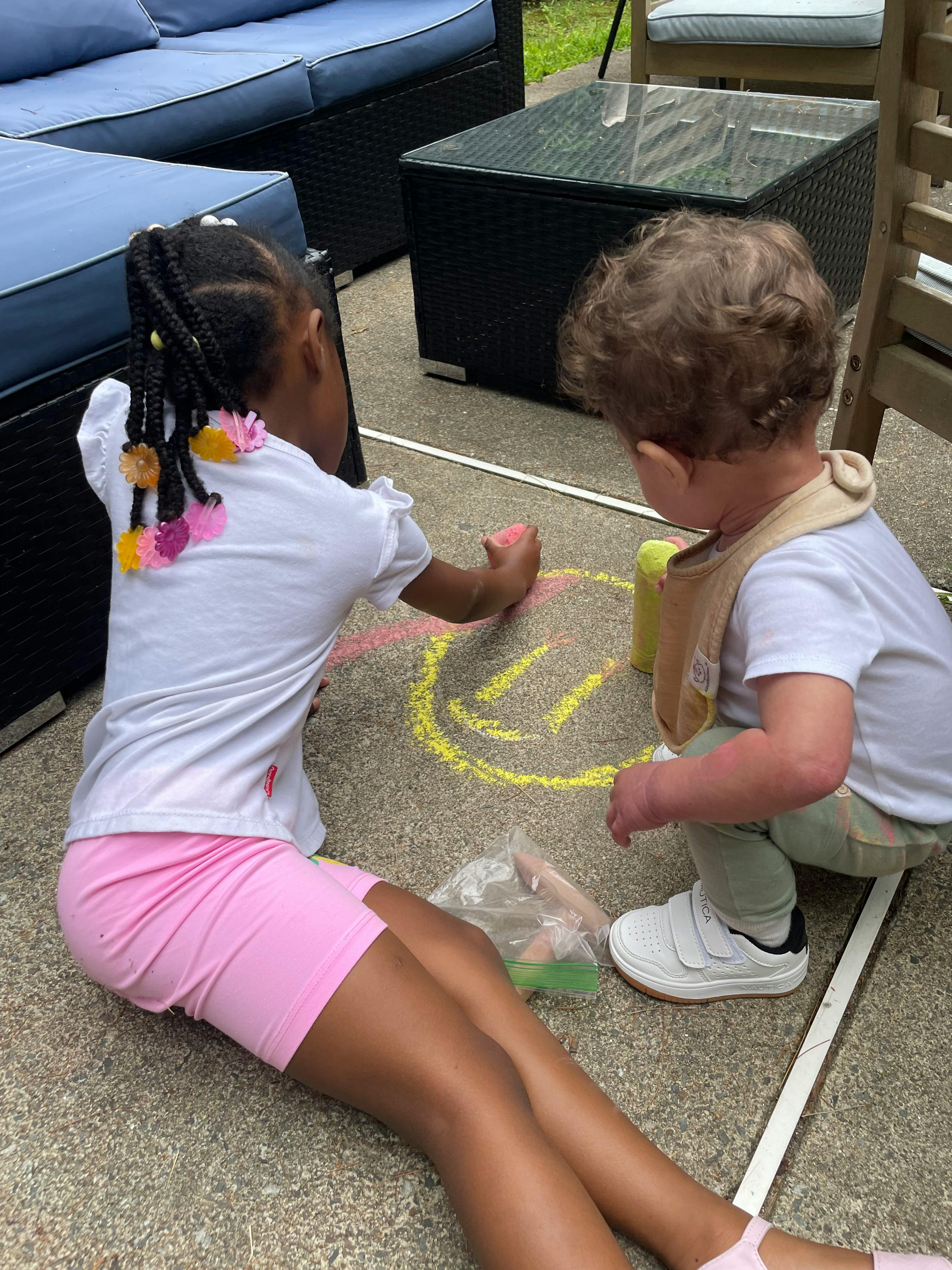 Child drawing a yellow smiley face with chalk on the ground, another child watching