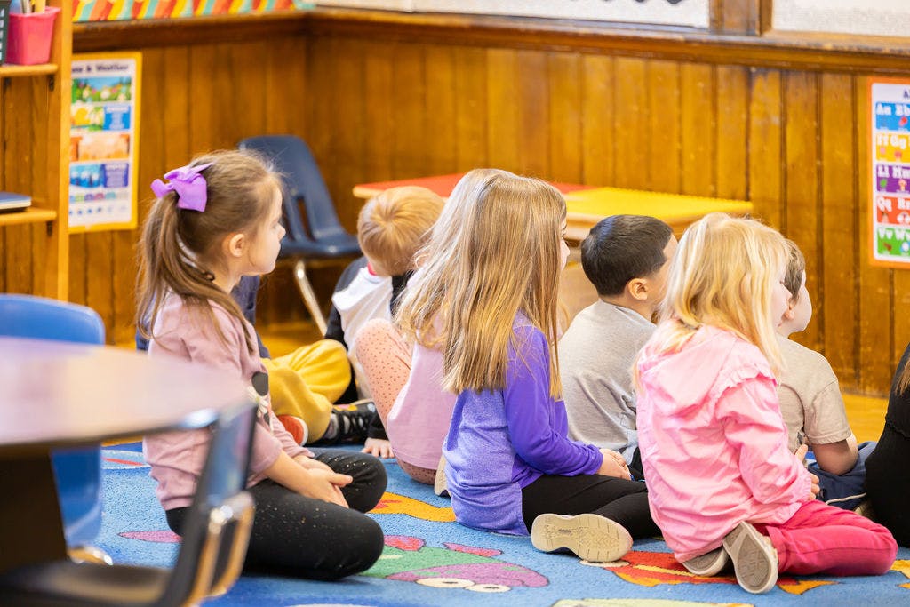 Children sitting on a rug in a classroom, viewed from behind and side