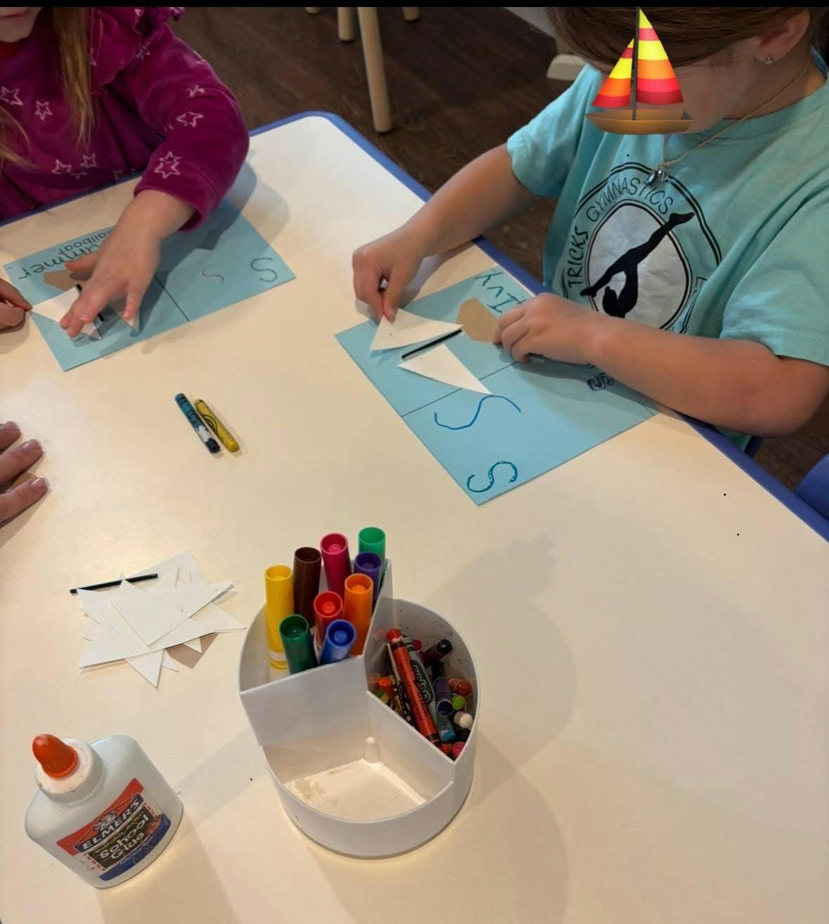 two children at a table doing a craft with paper, glue, and markers