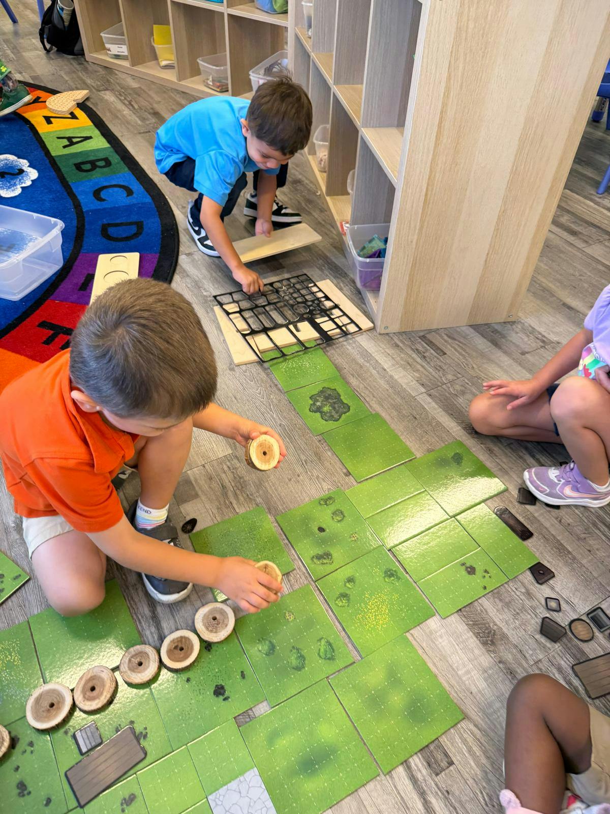 Children playing with green tiles and wooden pieces on the floor