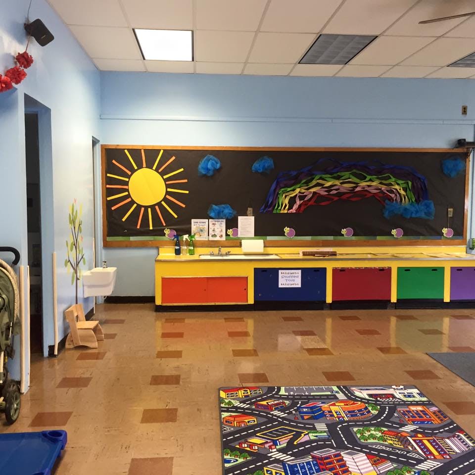 Classroom with a sun and rainbow bulletin board, colorful cabinets, and a play rug