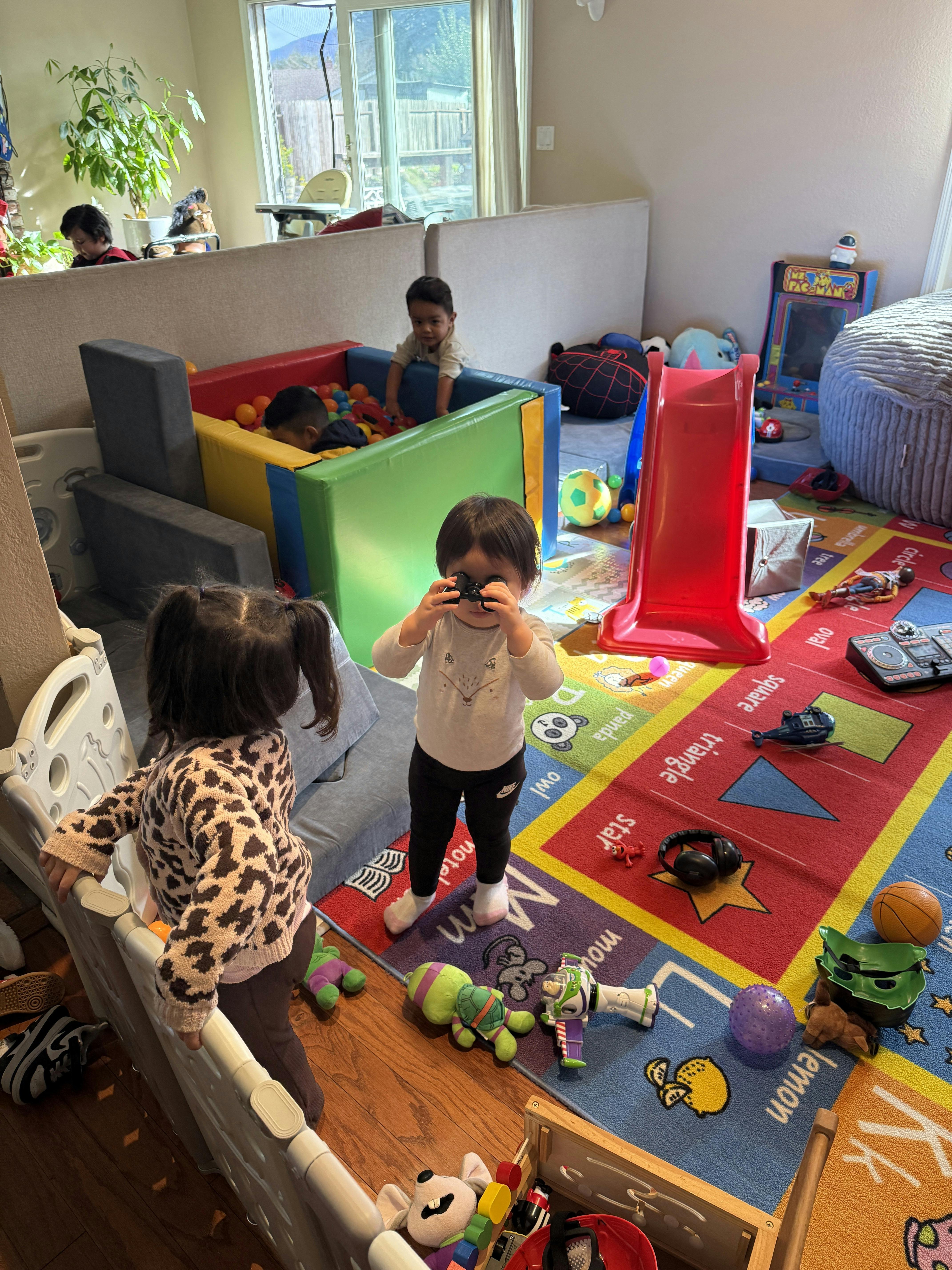 Child looking through binoculars while another child stands by a playpen