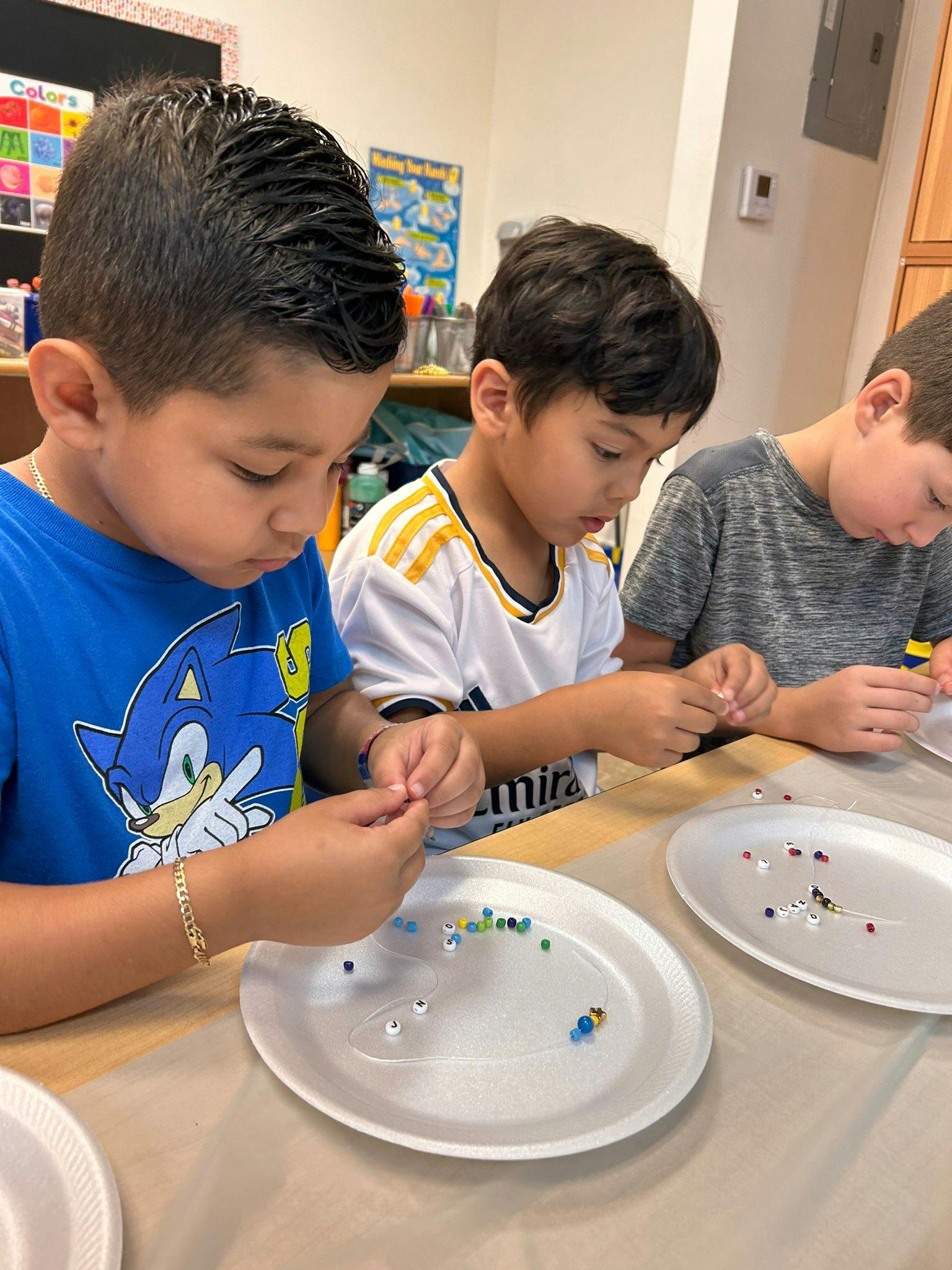 Three children sitting at a table stringing beads onto a thread