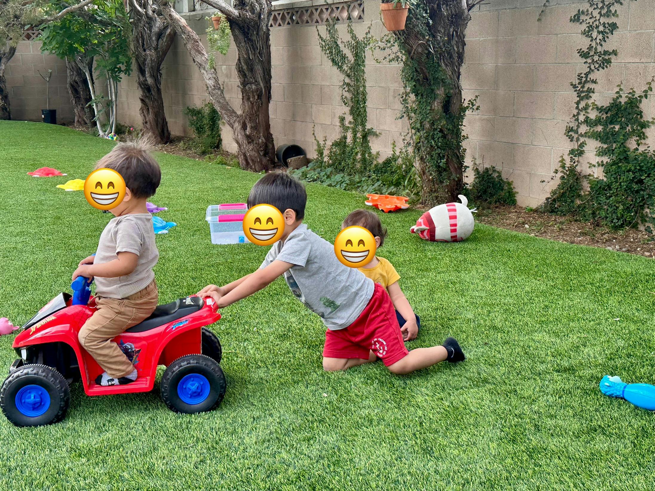 Child on a red toy ATV being pulled by another child on grass