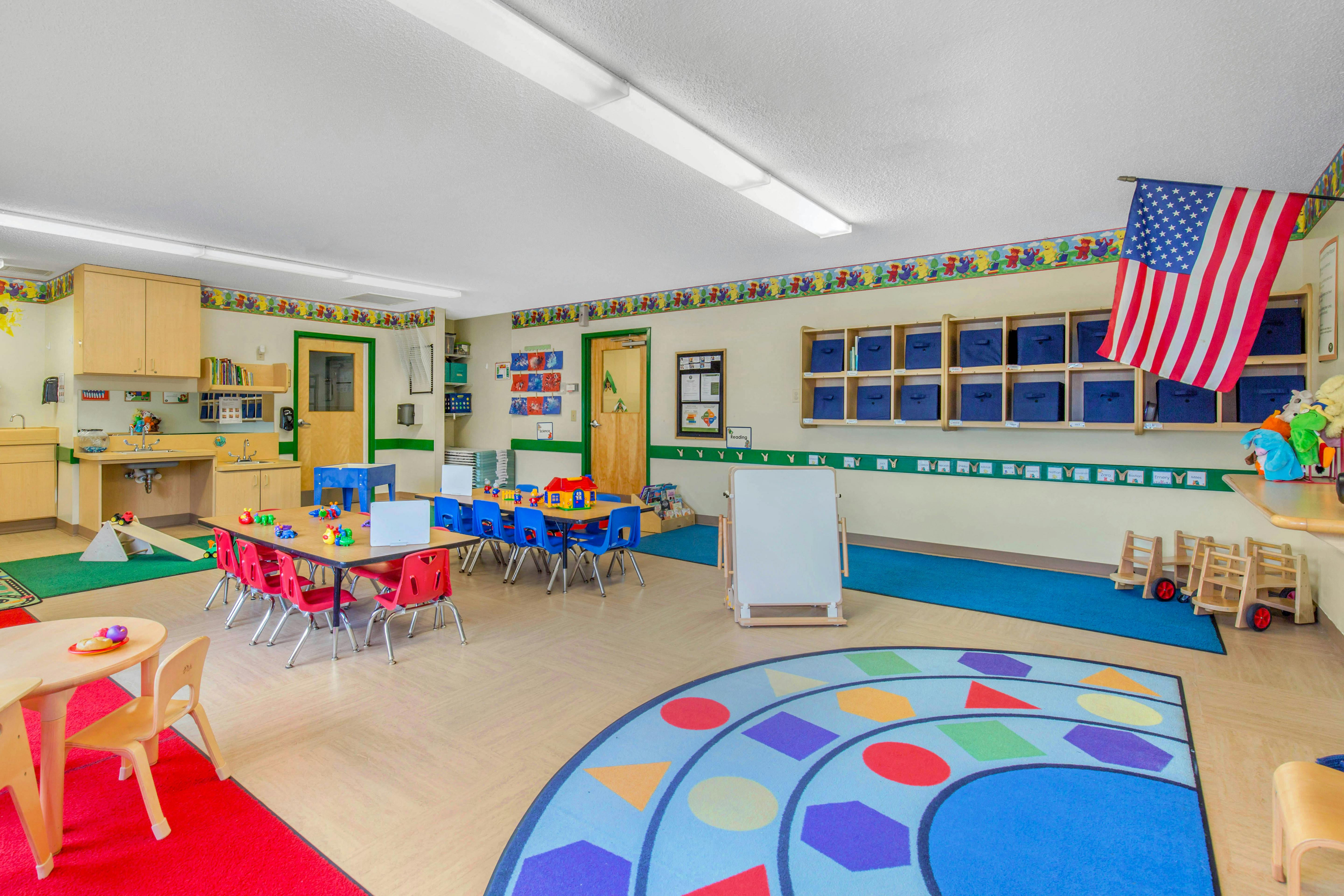 A classroom with small tables and chairs, a sink area, and toy storage shelves