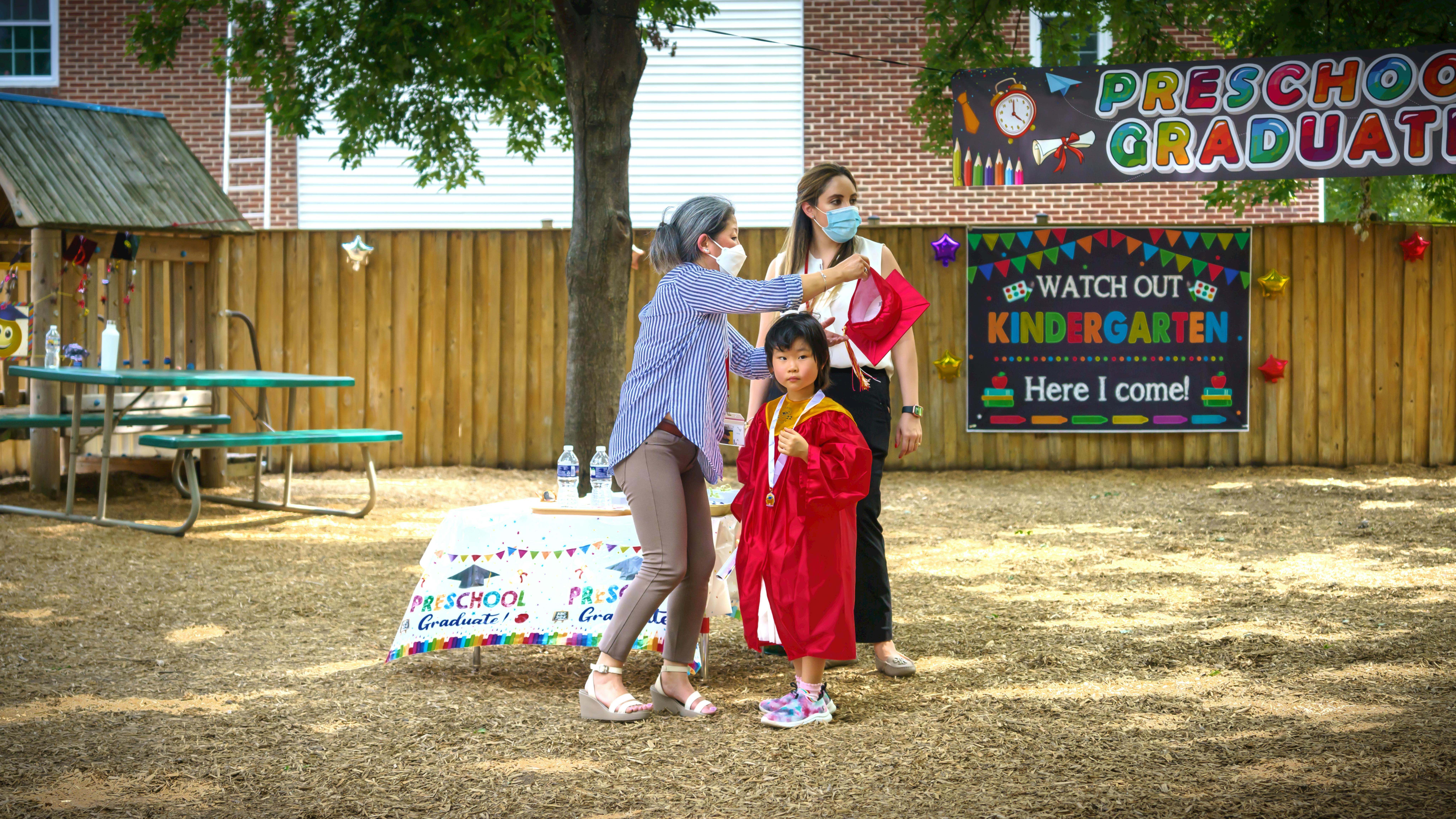 Adult assisting a child in a red graduation gown at a preschool ceremony