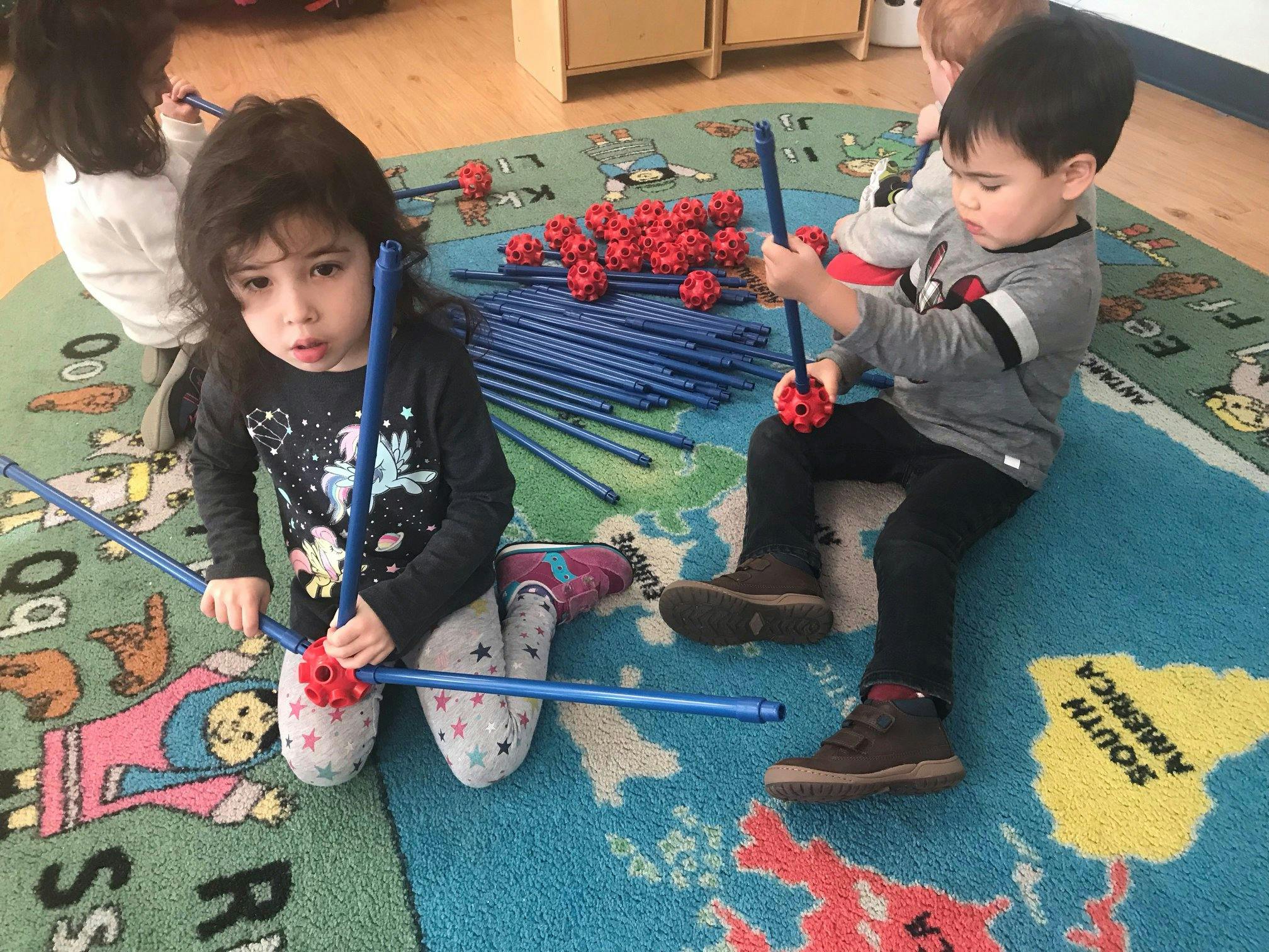 Two children playing with blue rods and red connectors on a map rug