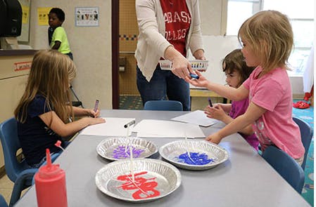 Adult handing a child a crayon at a table with other children and art supplies