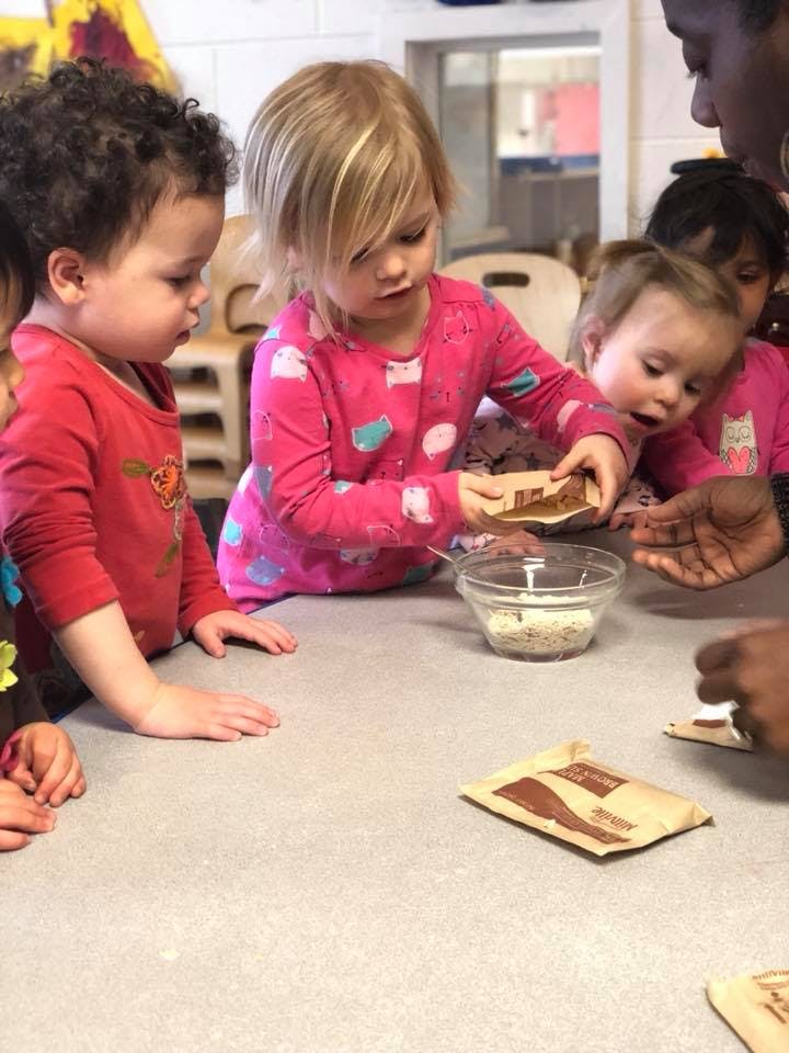 Children at a table with a person, one child holding a small package over a bowl