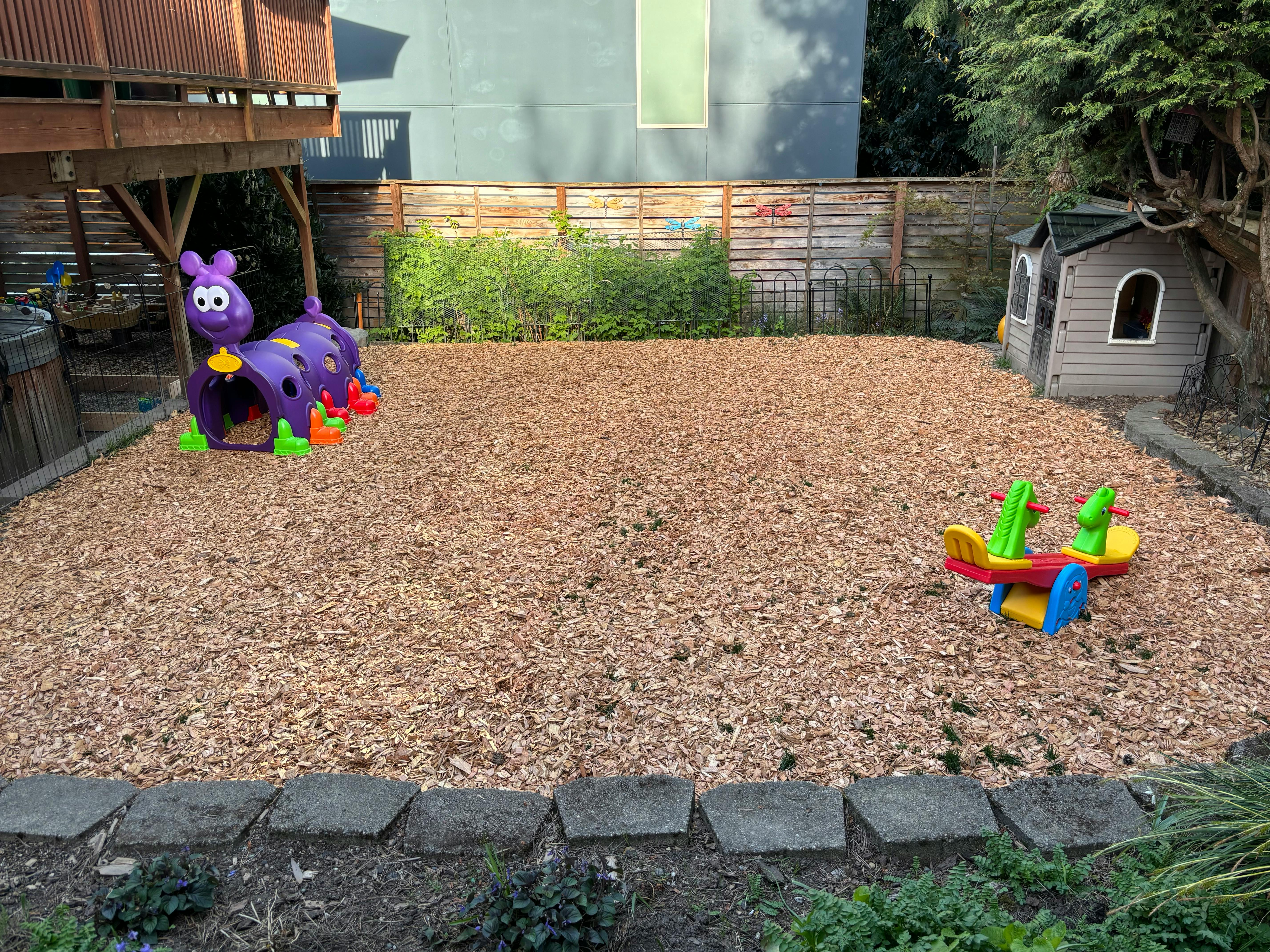 Backyard play area with wood chips, a purple caterpillar tunnel, and a toy plane